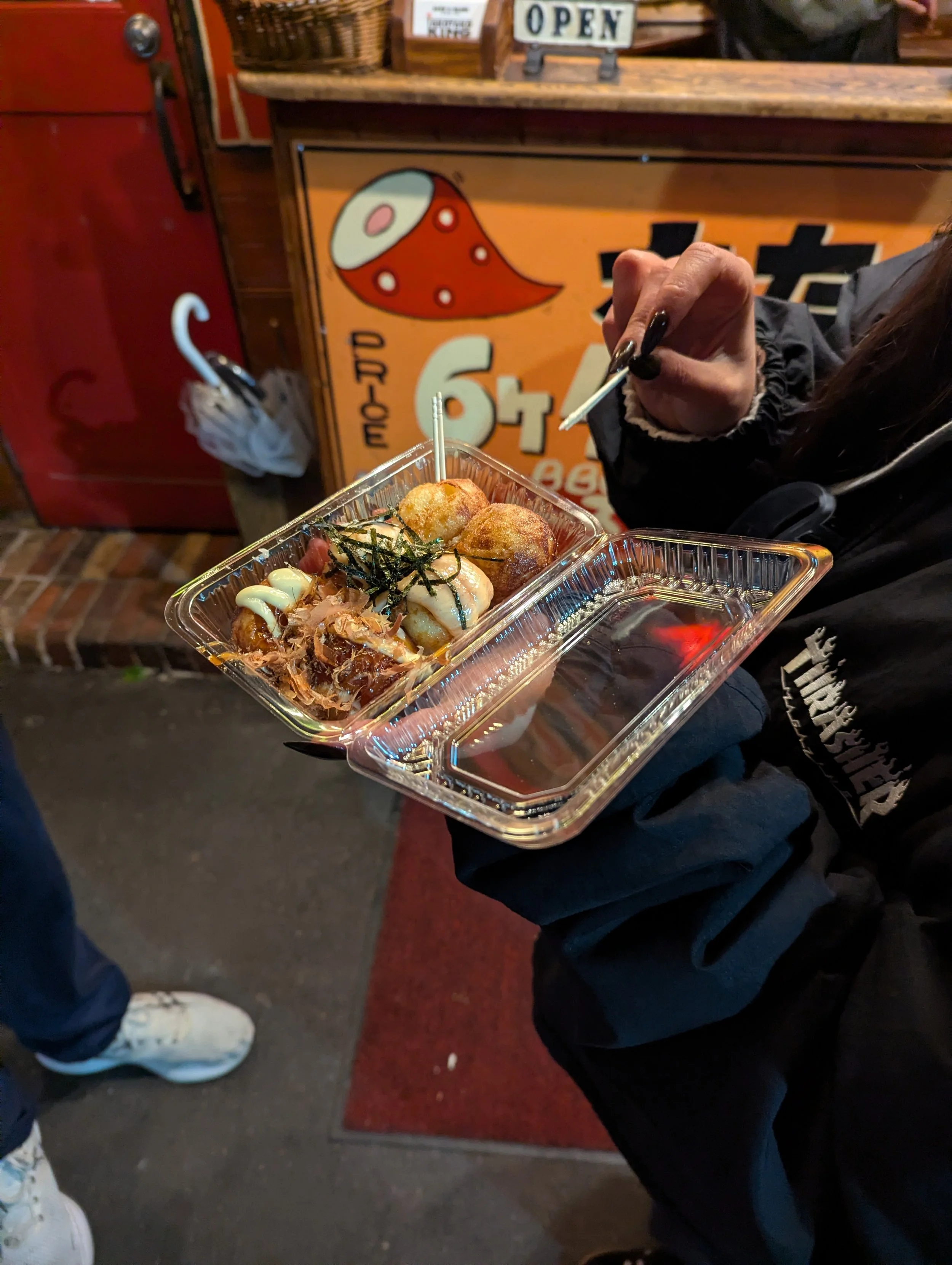 Person holding a plastic tray with takoyaki, a few pieces of seaweed, and dipping sauce outside a restaurant.