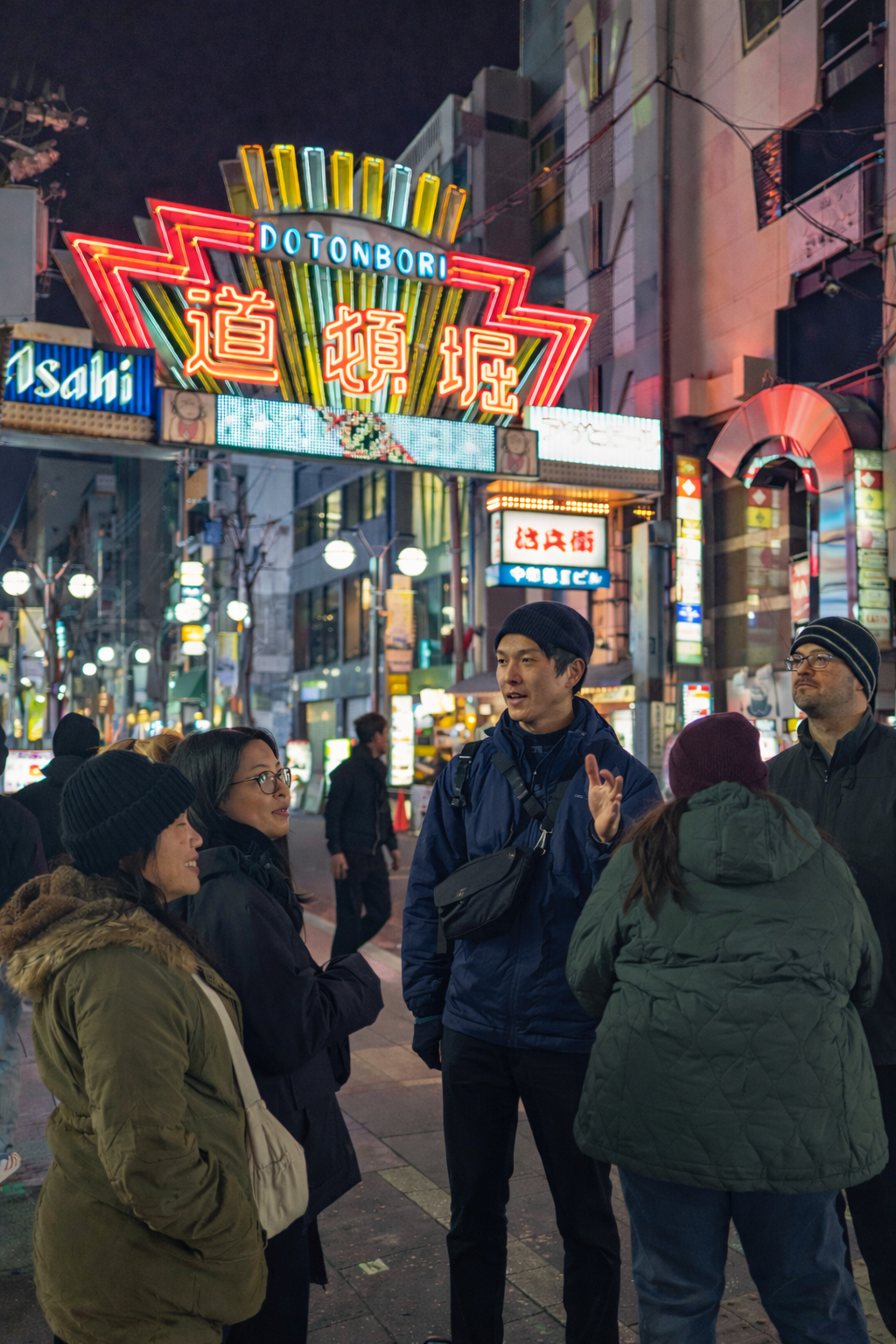 A group of five people chatting on a bustling street at night, illuminated by colorful neon signs in a city that appears to be in Japan.