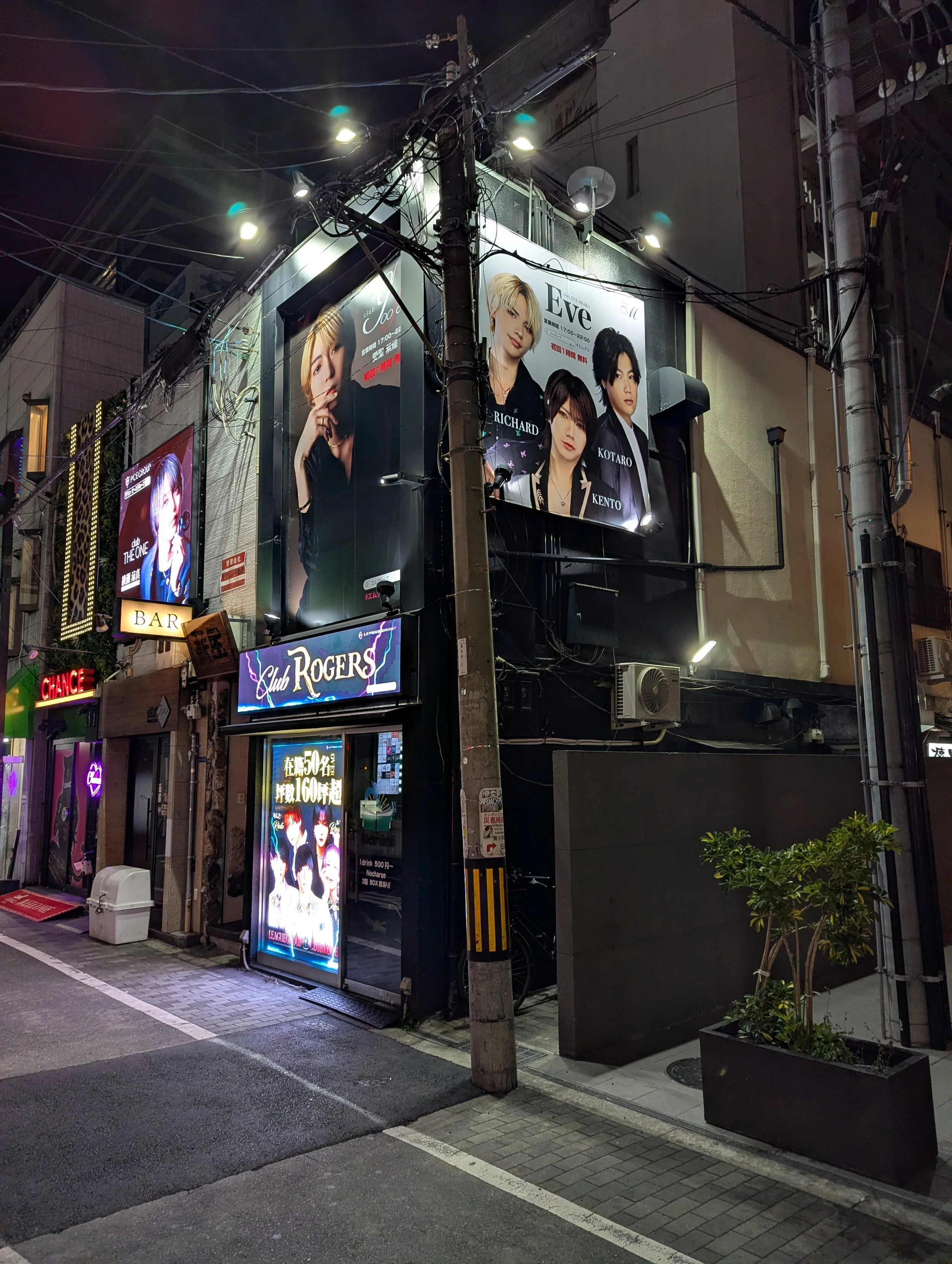 Night scene of a city street with illuminated signs and posters on buildings, including advertisements for a nightclub and a bar, with some greenery in planters and power lines overhead.