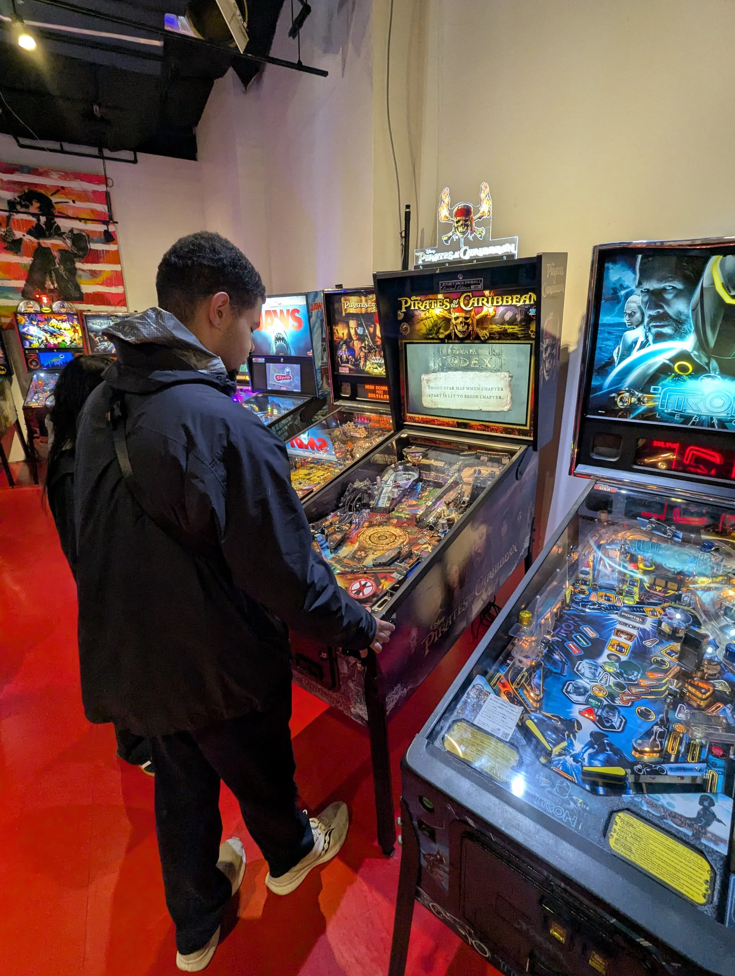 A young man in a black jacket and beige sneakers playing a pinball machine in an arcade with multiple pinball and arcade machines around him, including a 'Pirates of the Caribbean' pinball machine in front of him.