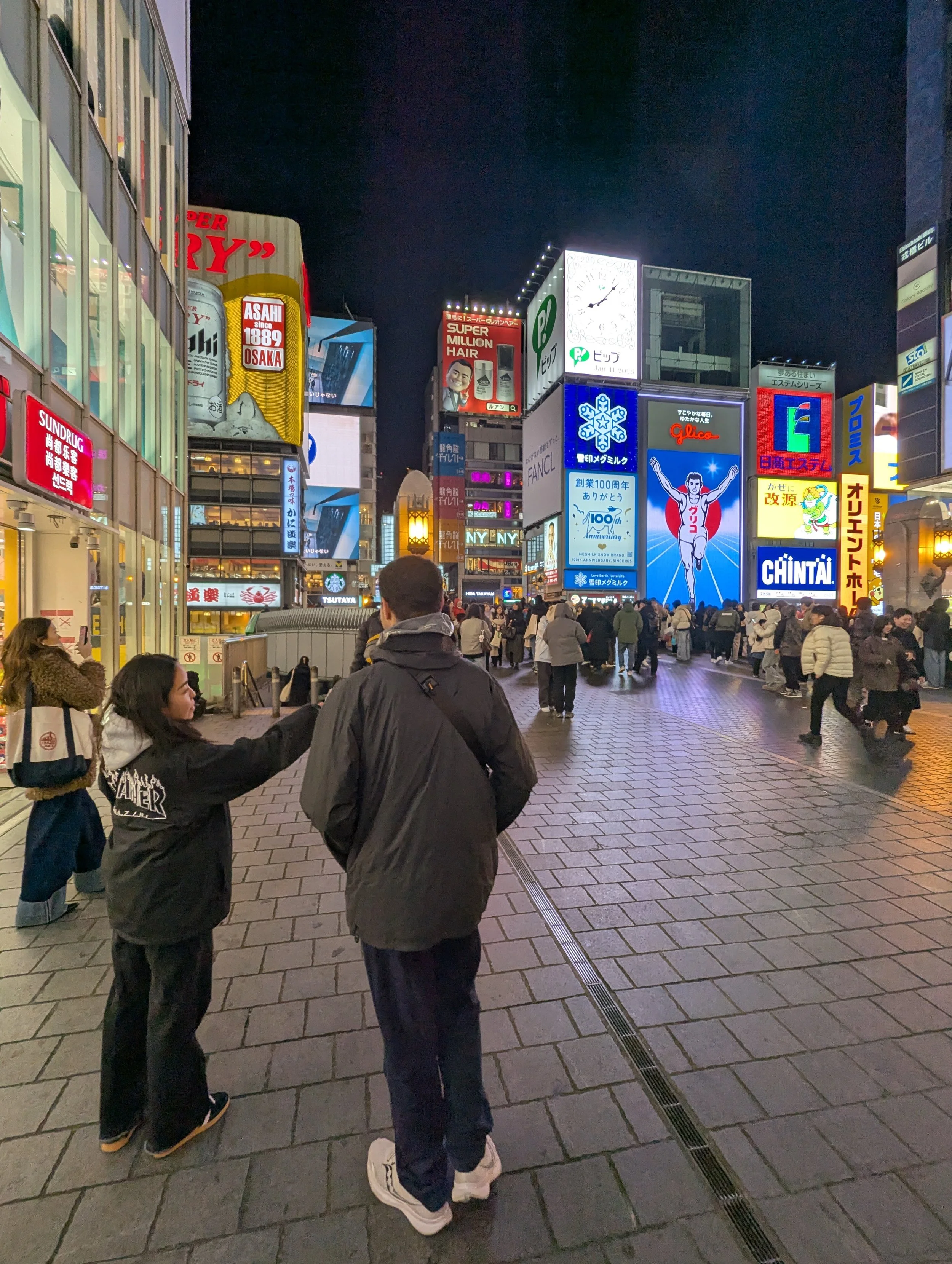 A busy city street at night with a crowd of people walking past brightly lit billboards and signs. Two people are in the foreground, with one talking to the other. The scene is vibrant with neon lights and advertisements.