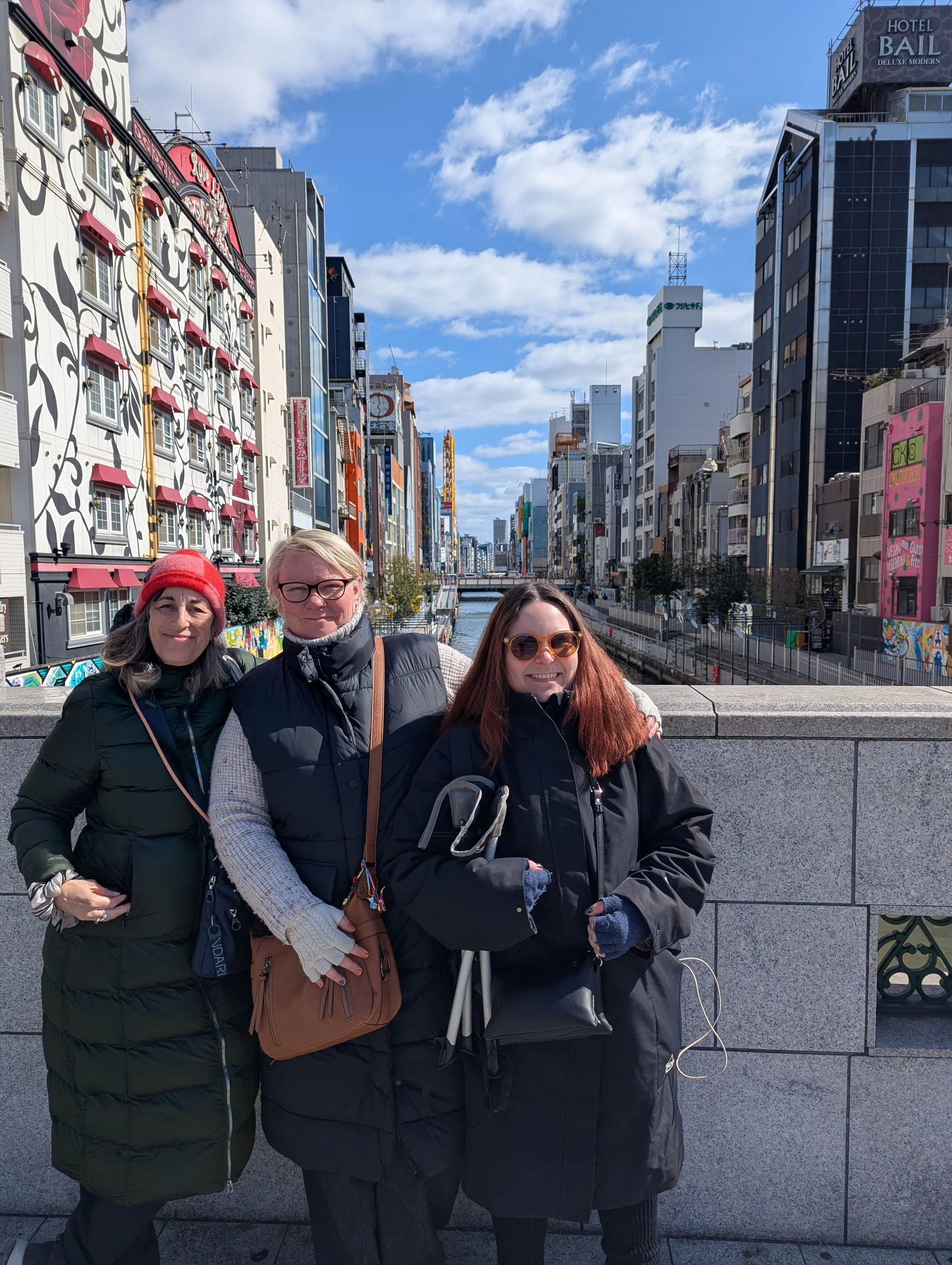 Three women standing on a bridge over a canal in an urban area, smiling at the camera. The background features colorful, modern buildings and a partly cloudy sky.