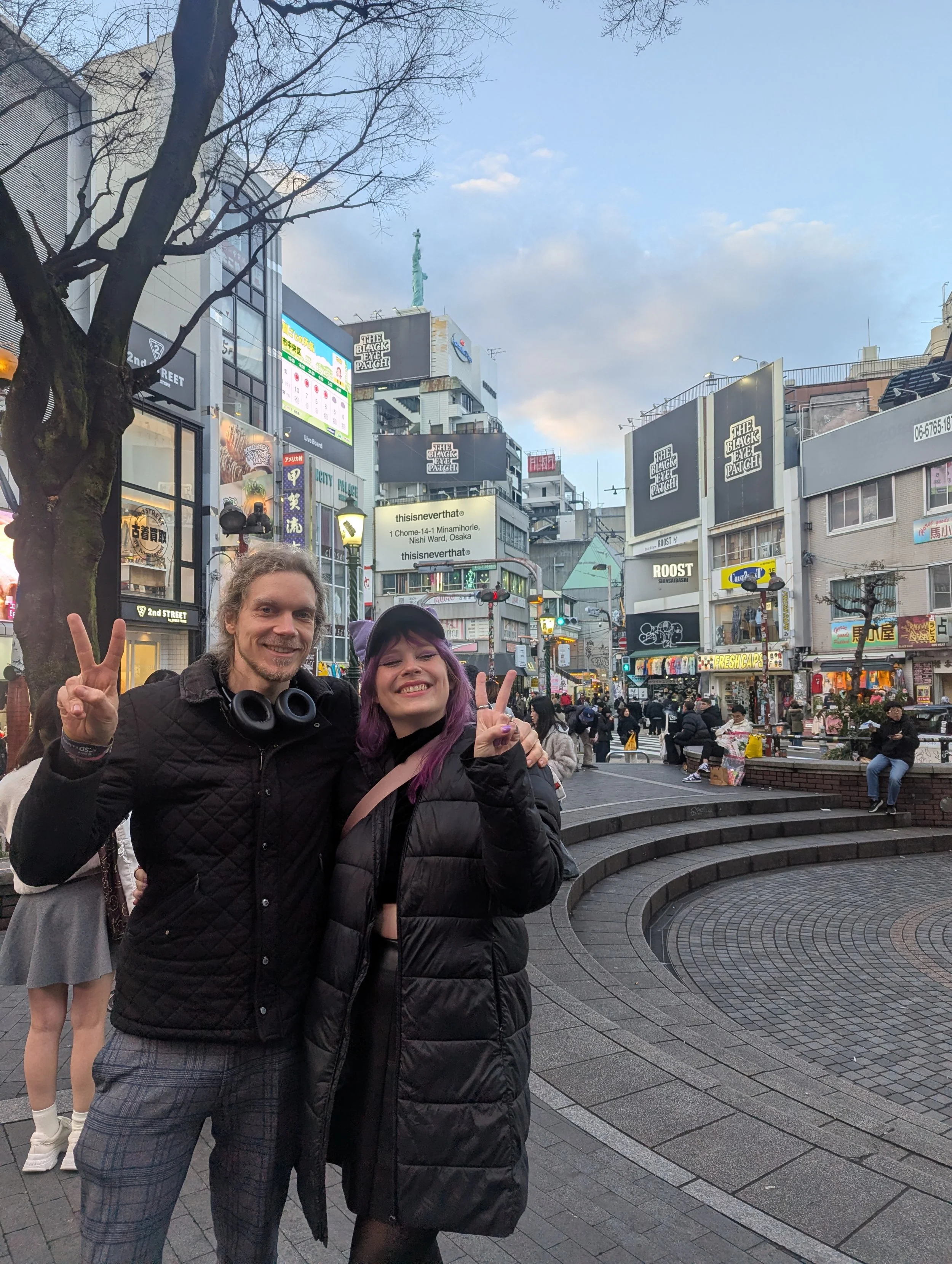 Two smiling people, a man and a woman, standing together on a city street in Japan, making peace signs with their hands. They are dressed warmly, with the woman sporting purple hair, against a backdrop of buildings, billboards, and pedestrians in an 