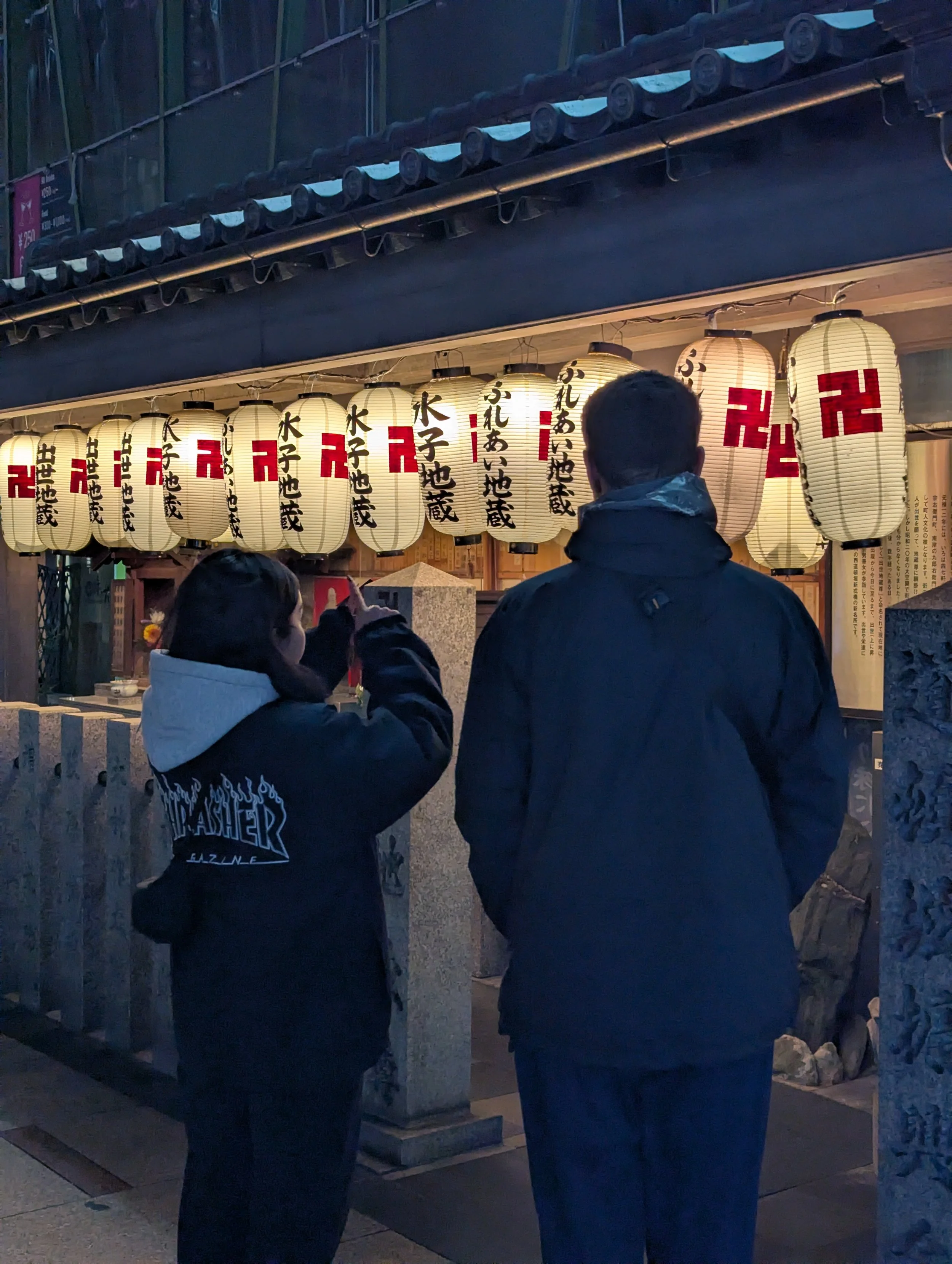 People standing in front of a row of traditional Japanese paper lanterns with kanji characters, at dusk or evening.
