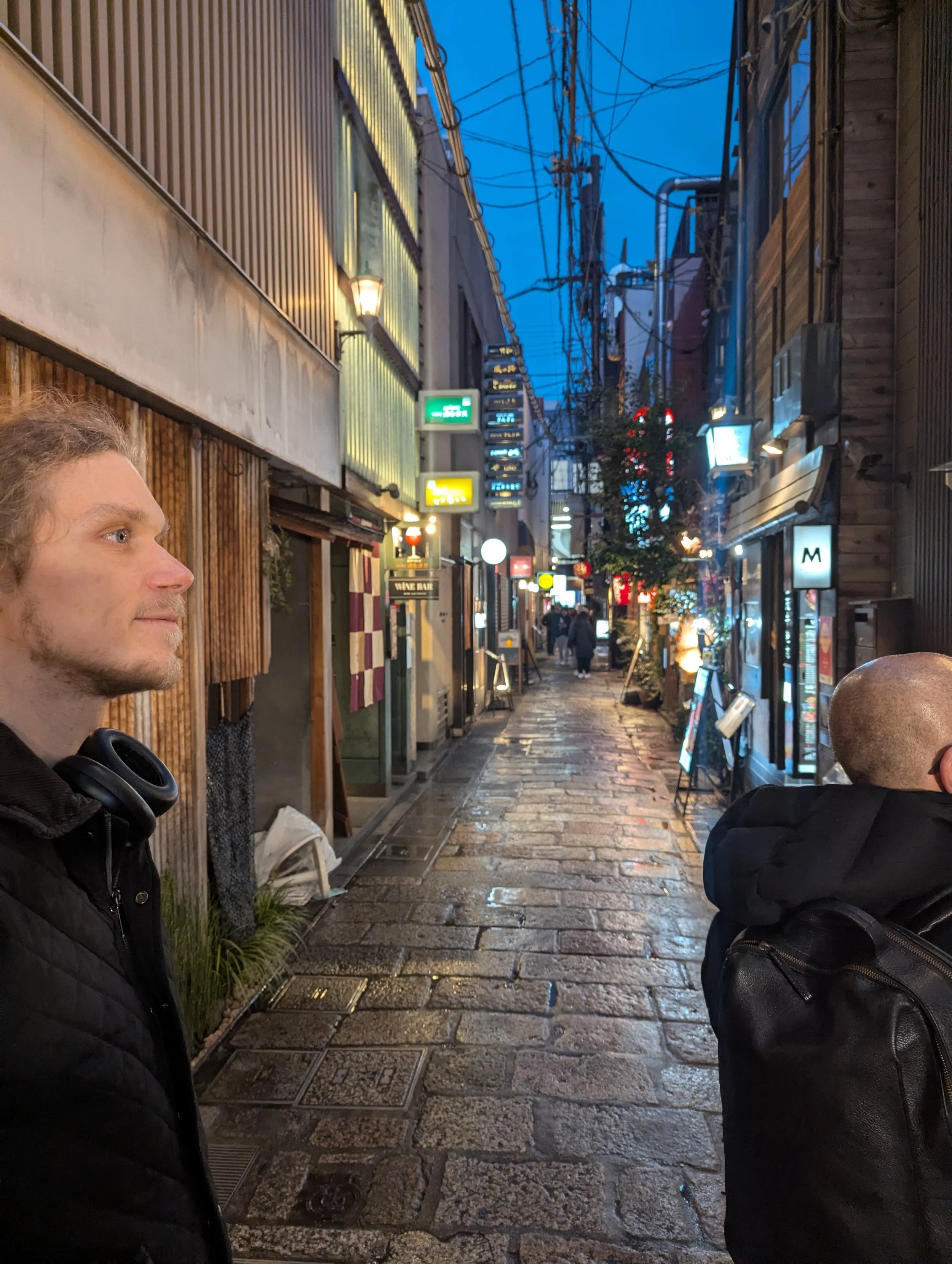 A narrow, wet cobblestone street in an urban area during the evening, lined with small shops and restaurants with illuminated signs, and a few pedestrians walking in the background. Two men are visible in the foreground, one on the left with a side p