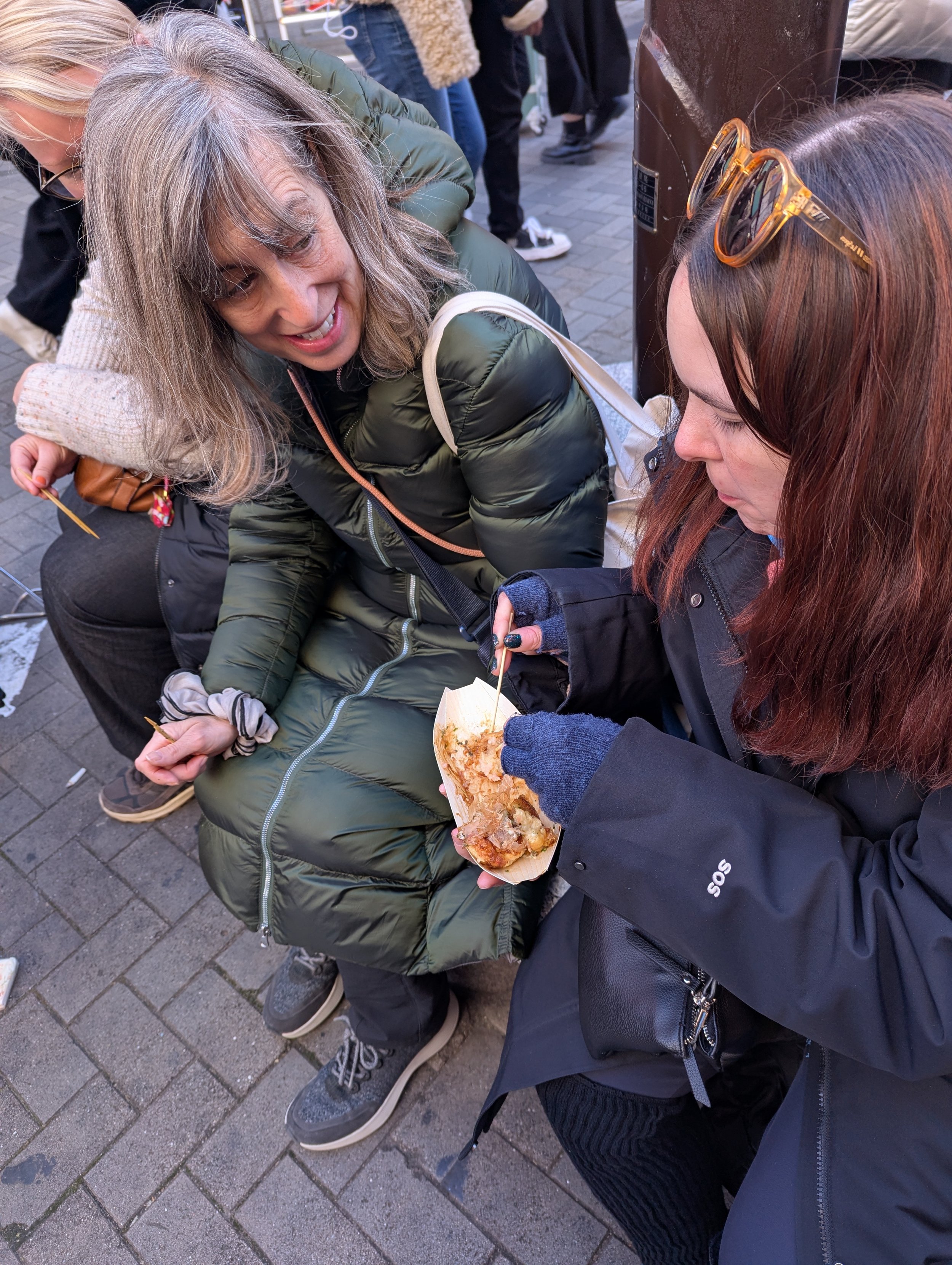 Two women sitting on a sidewalk; one with glasses and gray hair smiling, the other with red hair and sunglasses eating from a food tray.