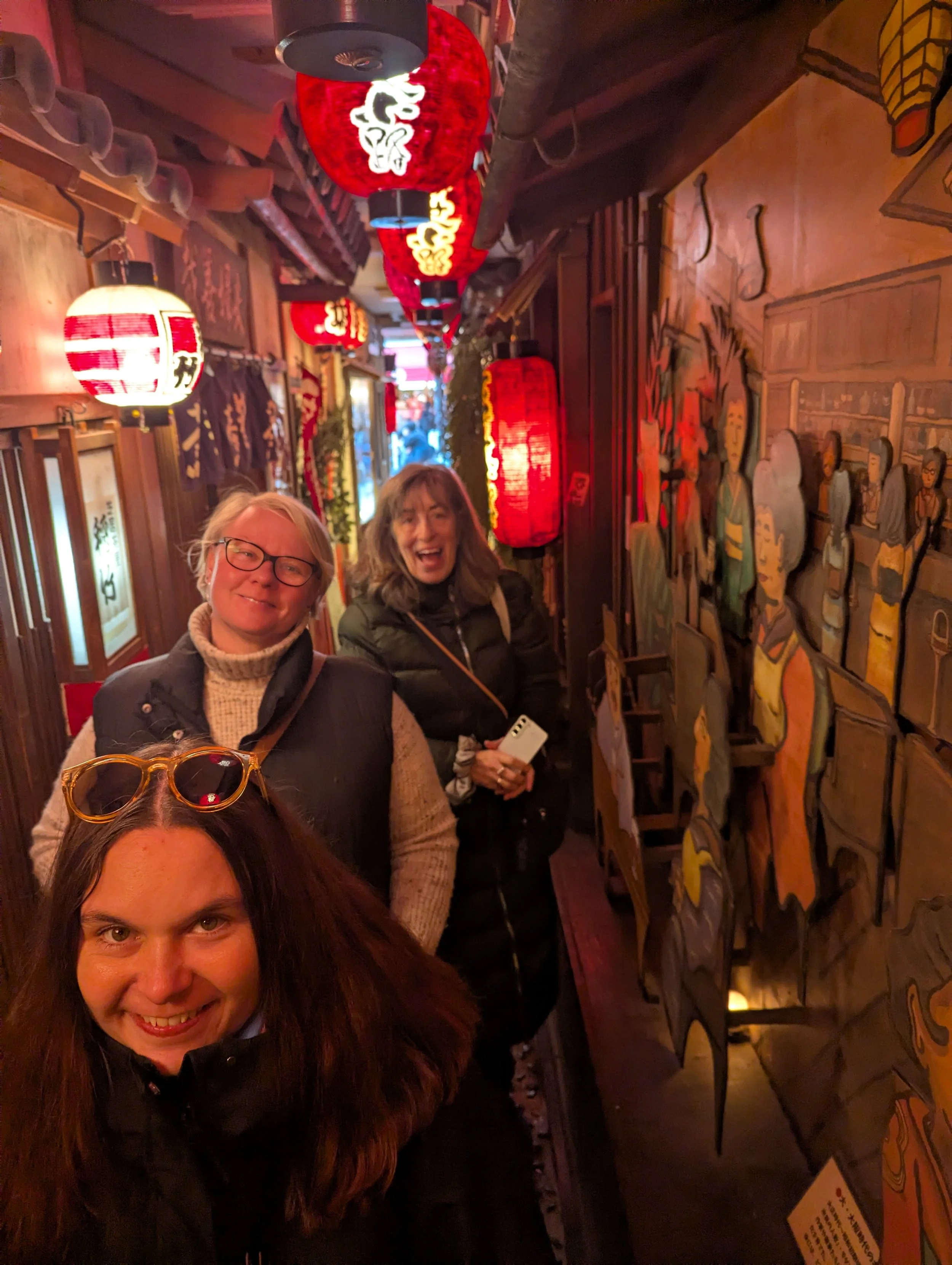 Three women smiling in a narrow,Japanese-themed restaurant decorated with red lanterns, traditional artwork, and wooden accents.