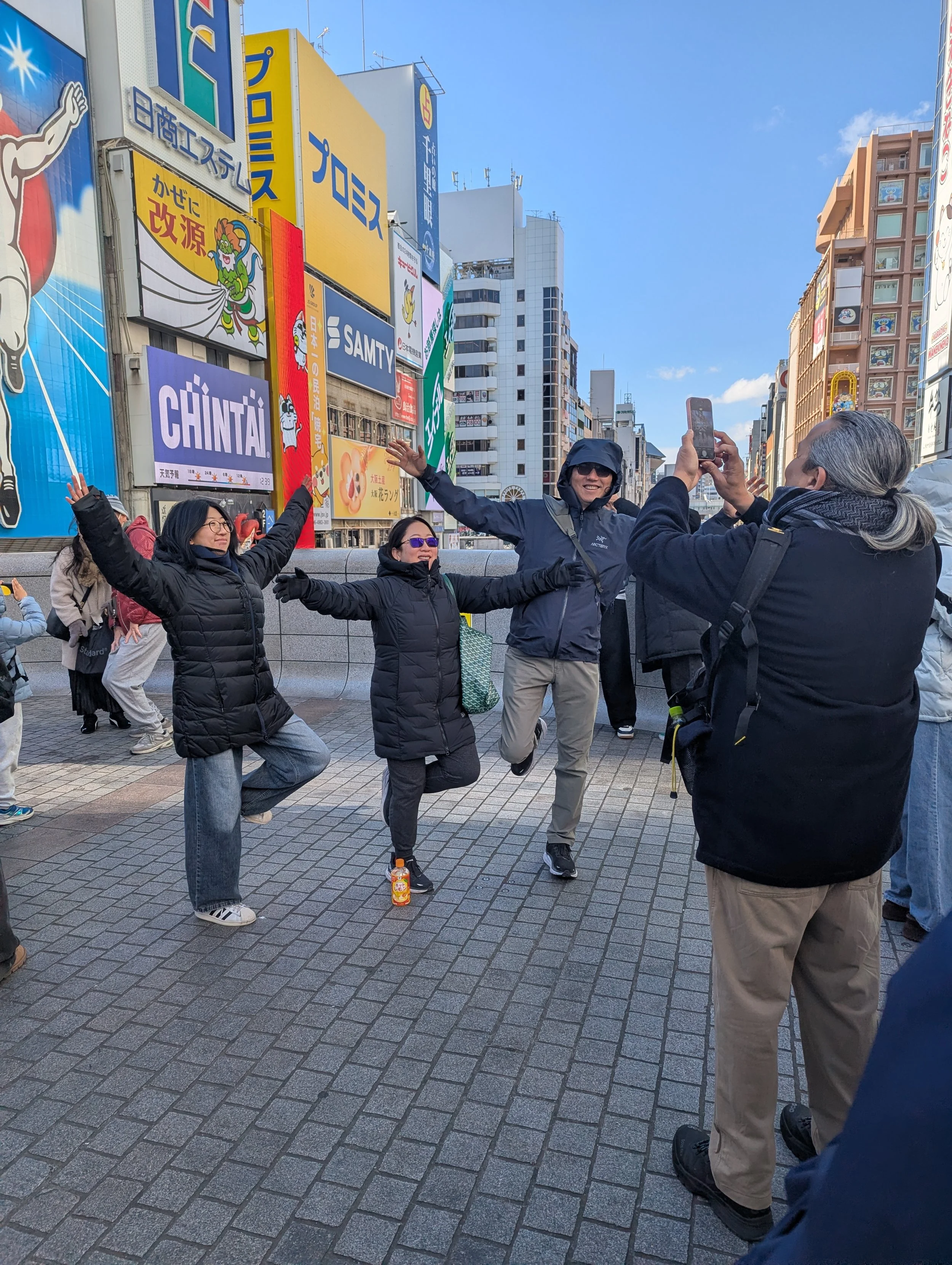 Four people standing on a city street, posing for a photo with arms outstretched and one person taking the picture. The background features colorful billboards and buildings, with a clear blue sky.