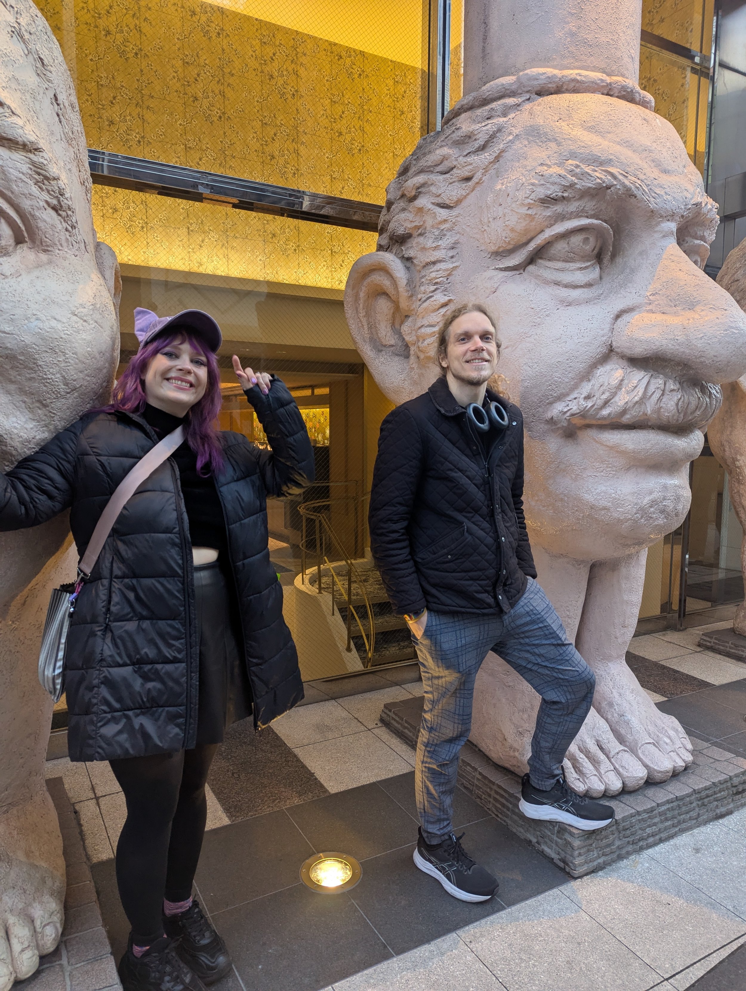 Two smiling people pose next to large stone sculptures of ancient-looking heads with prominent noses and mustaches outside a building with a yellow interior and glass windows.