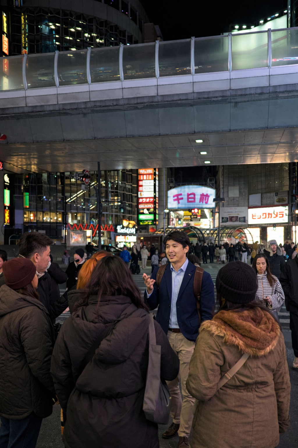 A group of people gathered outdoors in an urban area at night, with a cityscape and bright signs in the background, as a young man in a blue blazer talks to the crowd.