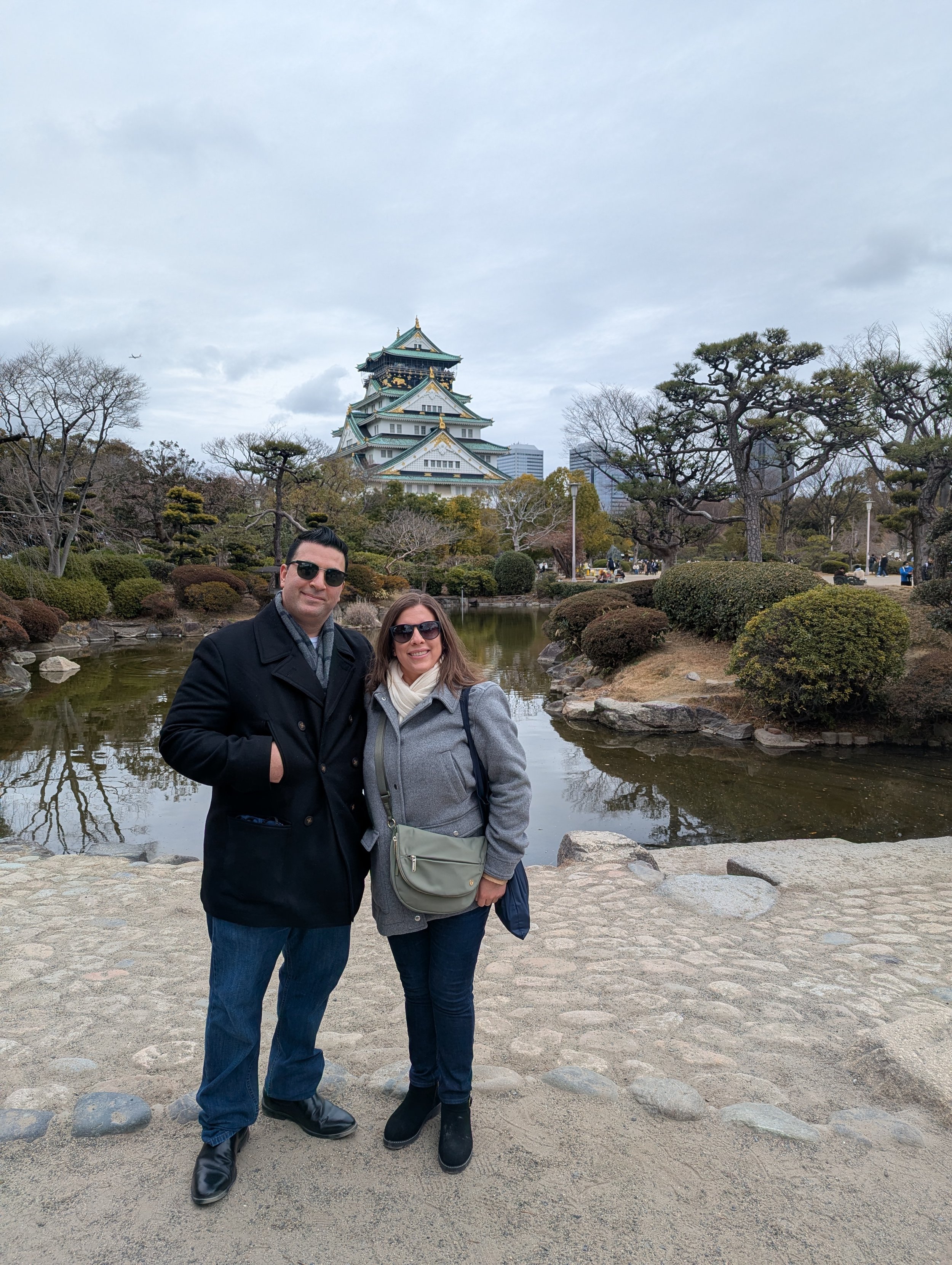 Two people standing on a stone path in a traditional Japanese garden with a pond, trimmed bushes, and trees, with a large multi-tiered castle in the background under cloudy skies.