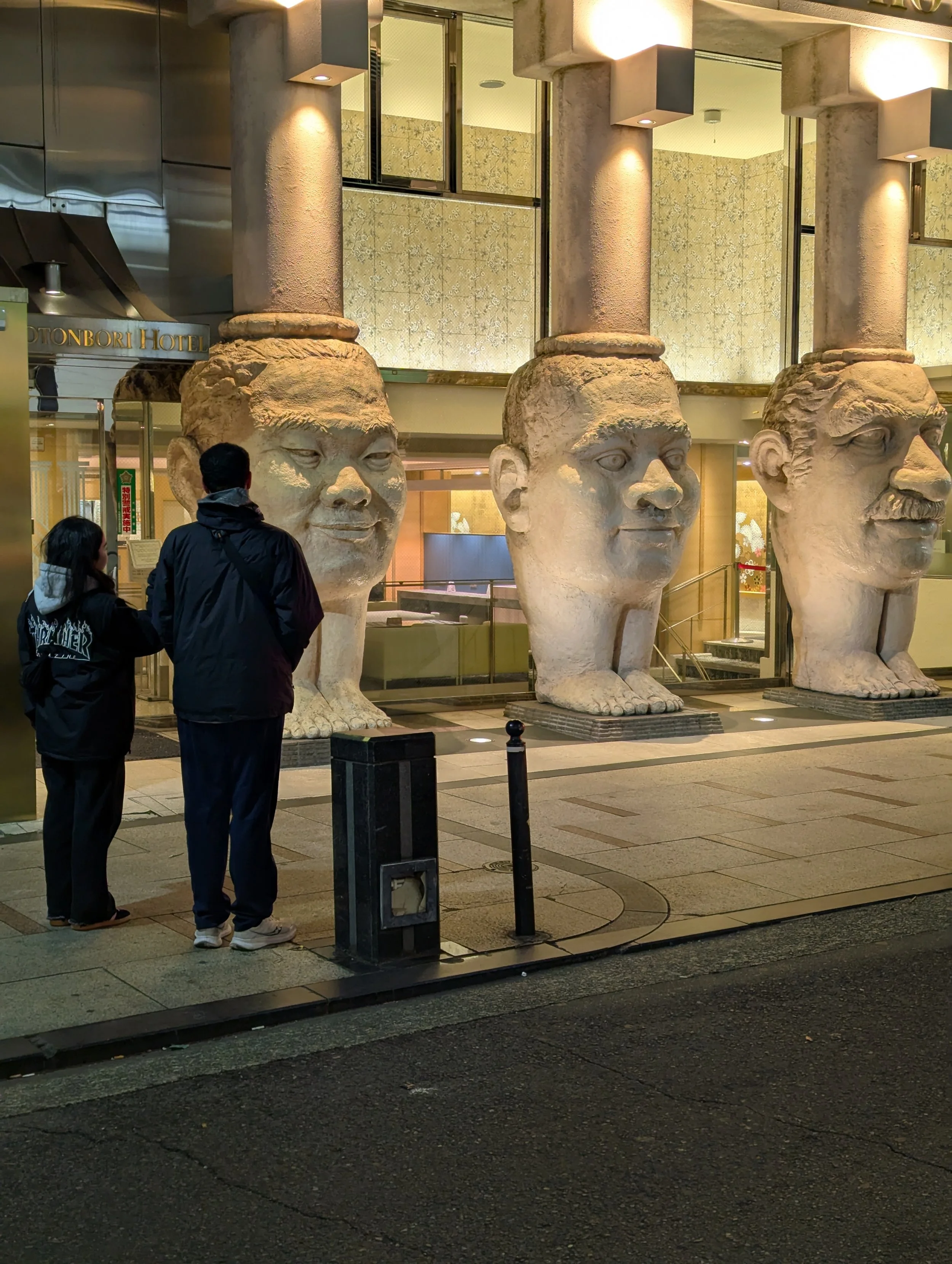 Three huge stone sculptures with human faces and large bodies with feet, positioned outside a building entrance, with two people standing nearby and talking.
