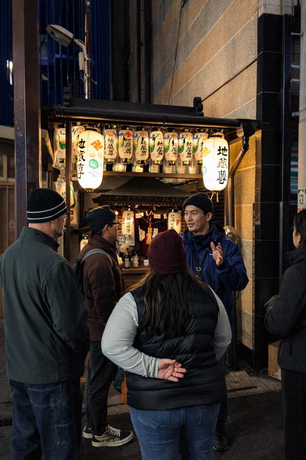A group of people gathered outside a small shop decorated with paper lanterns, some of which have Japanese characters on them. The shop appears to be in an urban setting at night.
