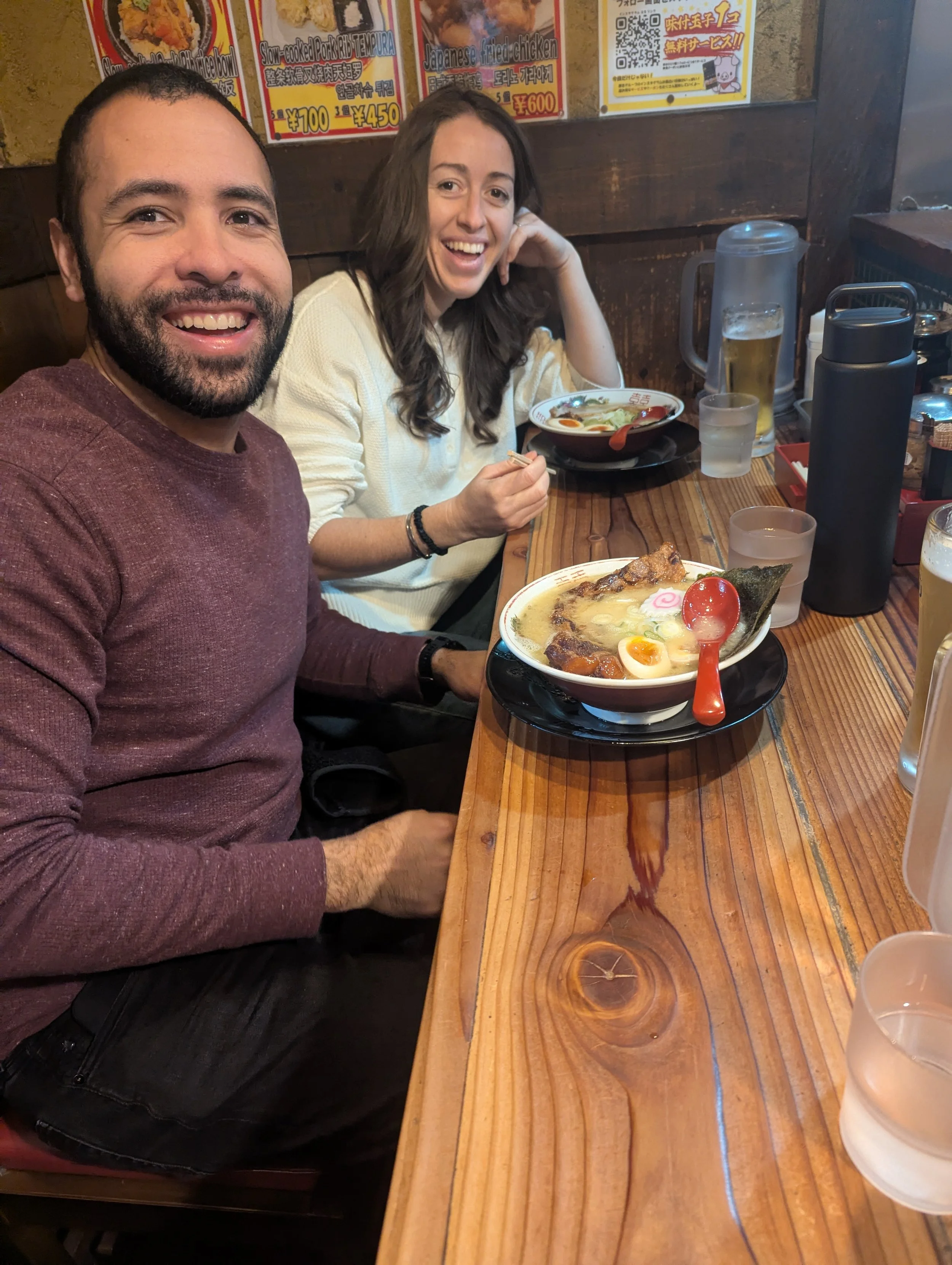 Two smiling people, a man and a woman, sitting at a wooden table in a restaurant with bowls of ramen in front of them. The woman is holding chopsticks, and there are drinks and condiments on the table.