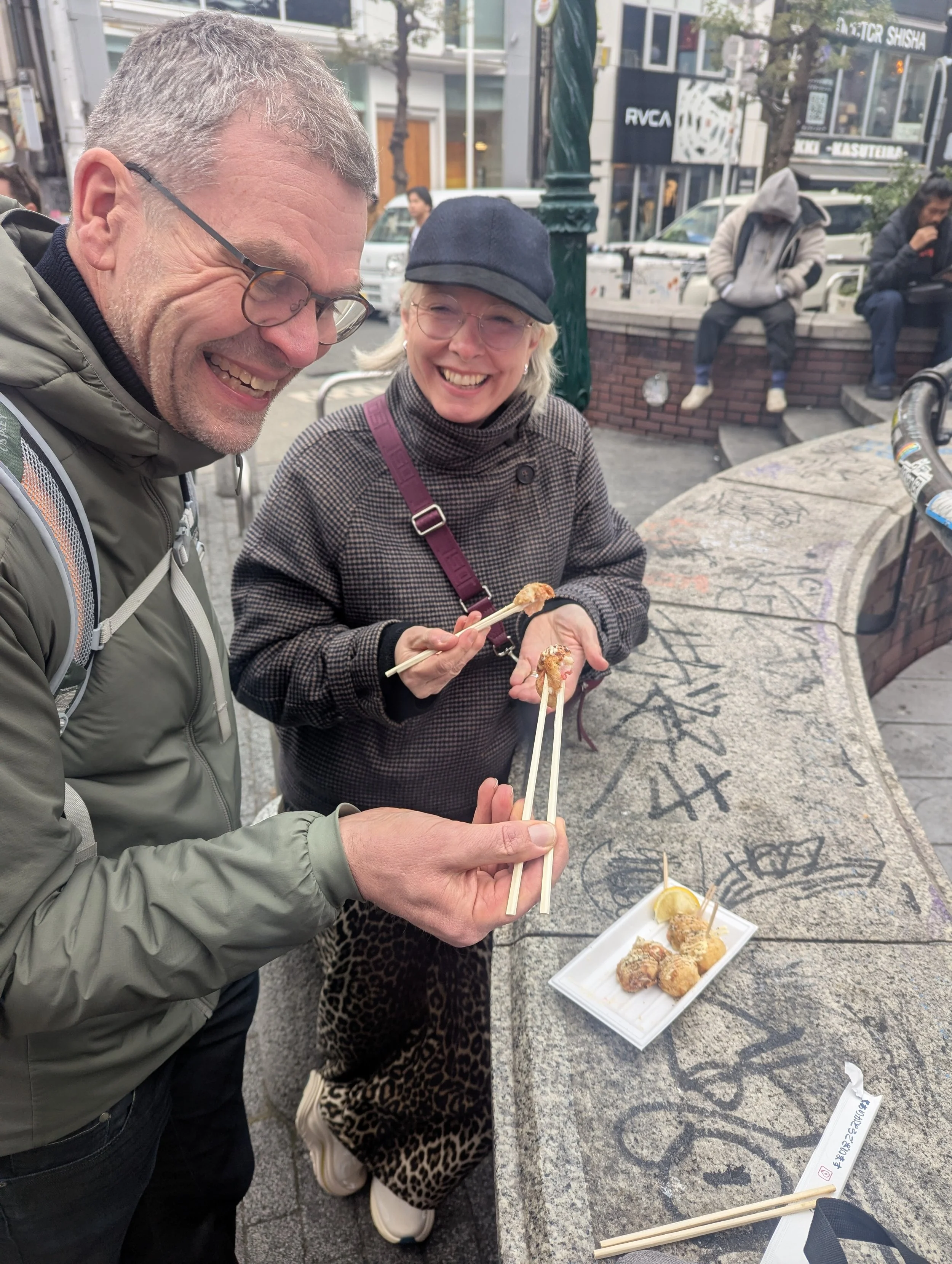 Two smiling people, a man and a woman, enjoying street food with chopsticks, standing beside a graffiti-covered concrete bench in an urban area.