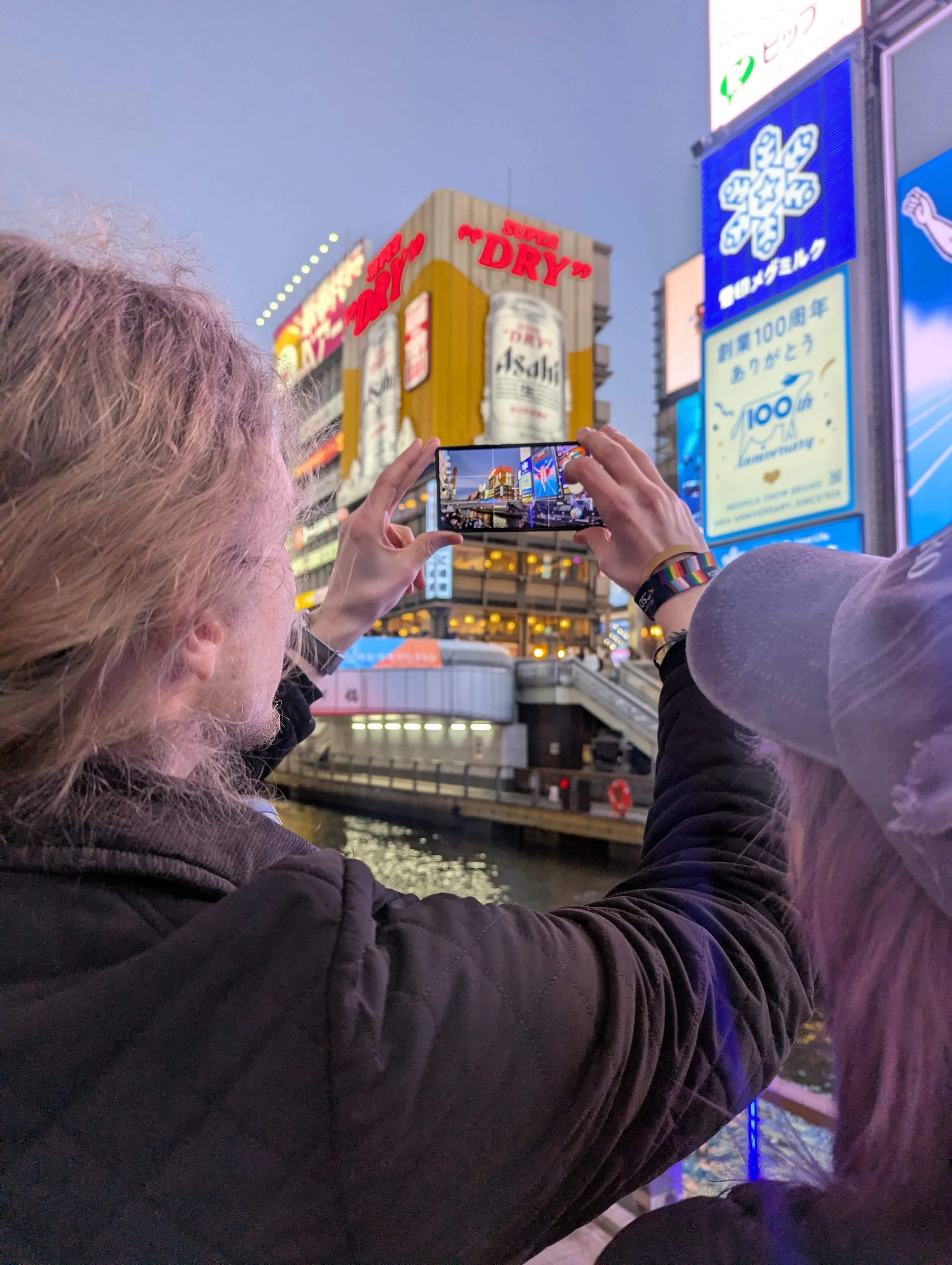 People taking a photo of bright, illuminated billboards and signs in Times Square, New York City during dusk.
