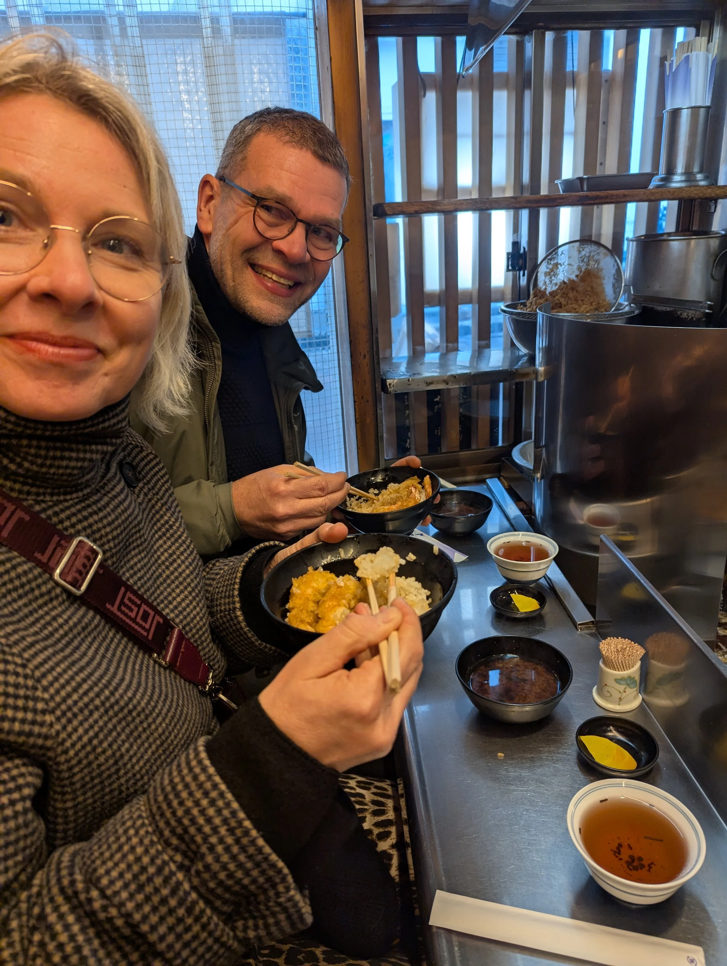 Two people are enjoying a meal at a restaurant counter, eating bowls of rice and tempura, with small bowls of tea and condiments on the counter.