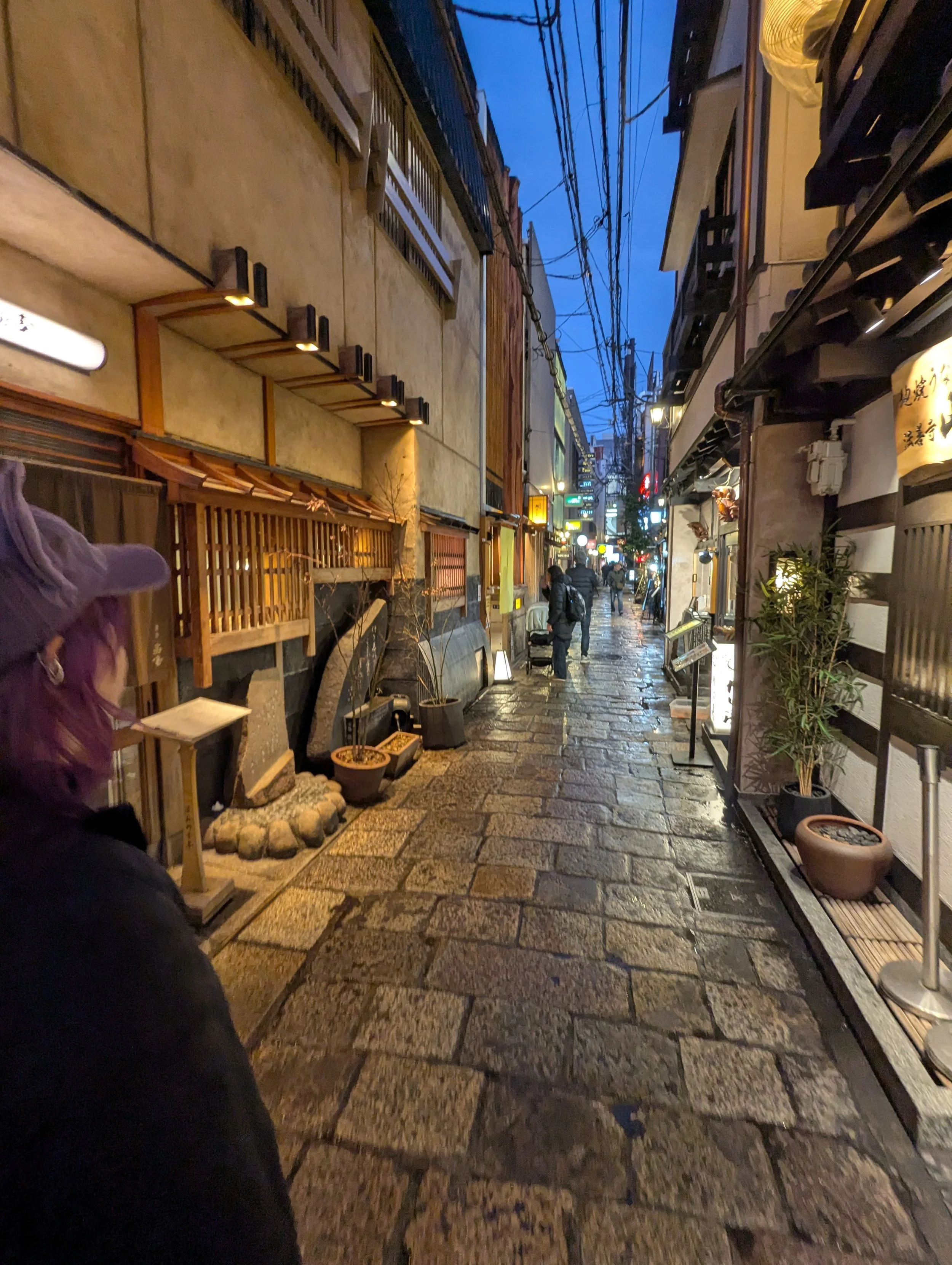 A narrow, wet street in Japan lined with traditional wooden buildings and lanterns, with a few people walking.