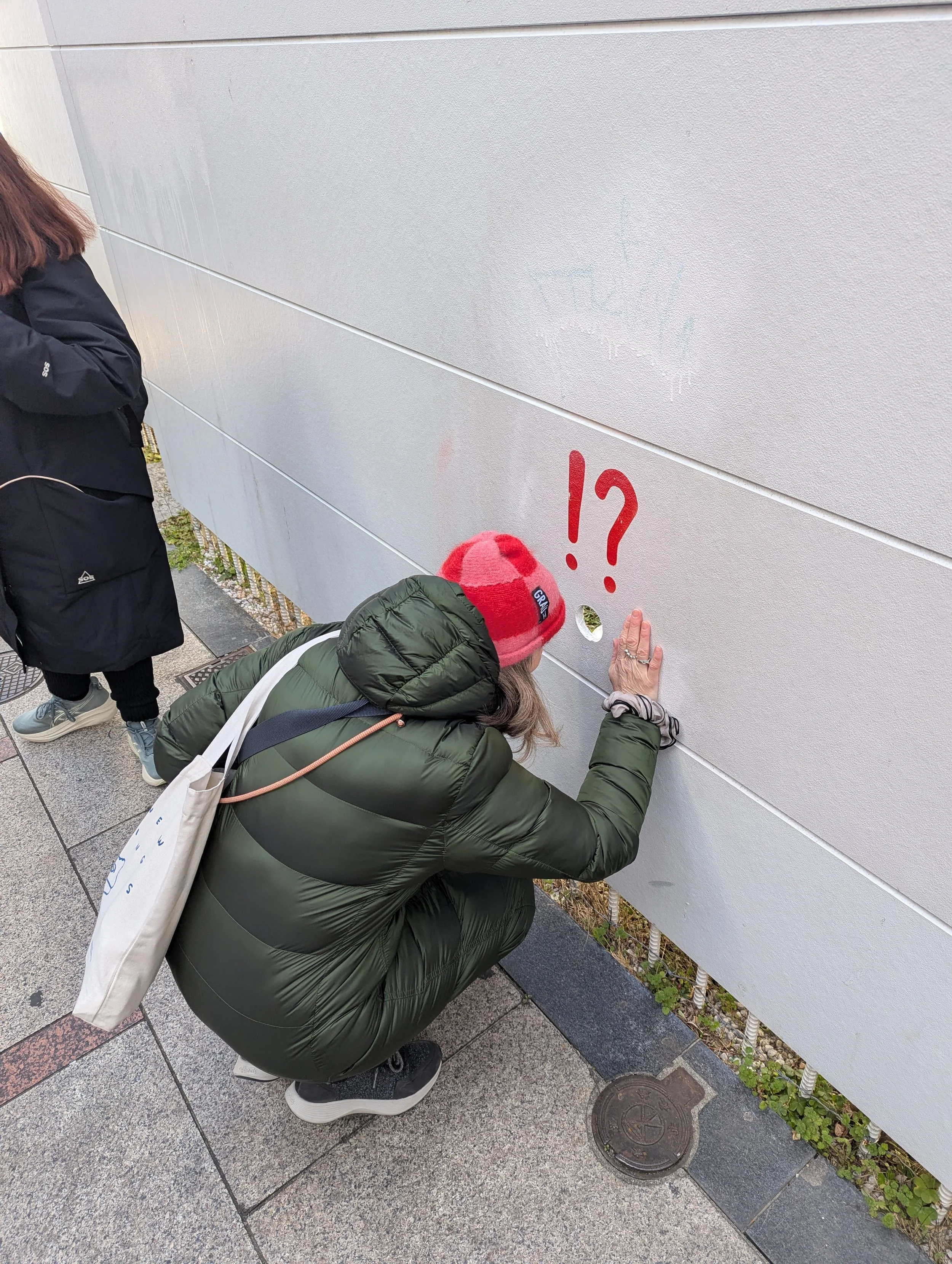 A woman squatting down and touching a hole in a wall with red question and exclamation marks painted above it. Another person stands nearby with a black jacket and backpack.