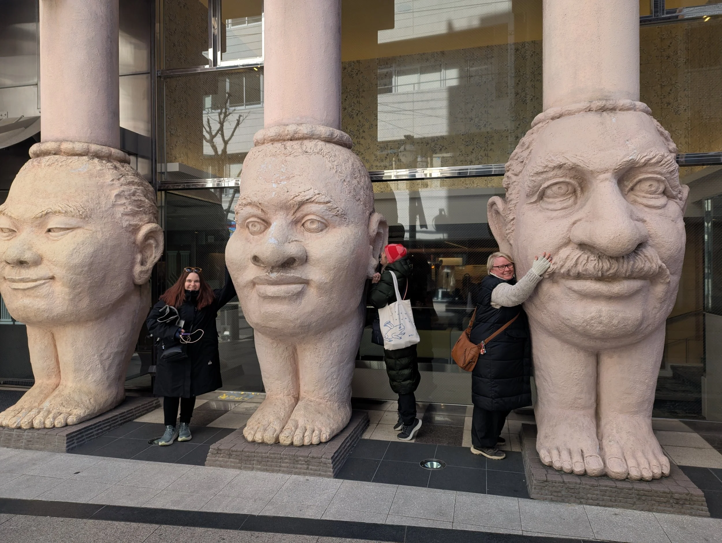 Three large sculptures of human heads with oversized feet in front of a building, with three women interacting with the sculptures, two hugging the heads and one standing nearby.