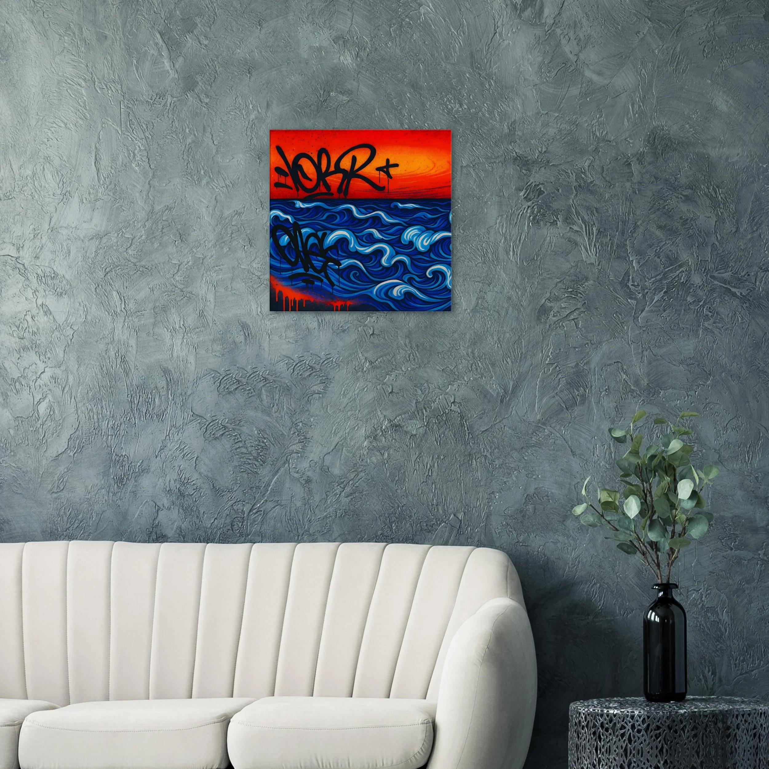 Wall-mounted colorful painting of ocean waves with graffiti text over a textured gray wall in a modern living room with a white sofa and a black vase with green leaves on a side table.