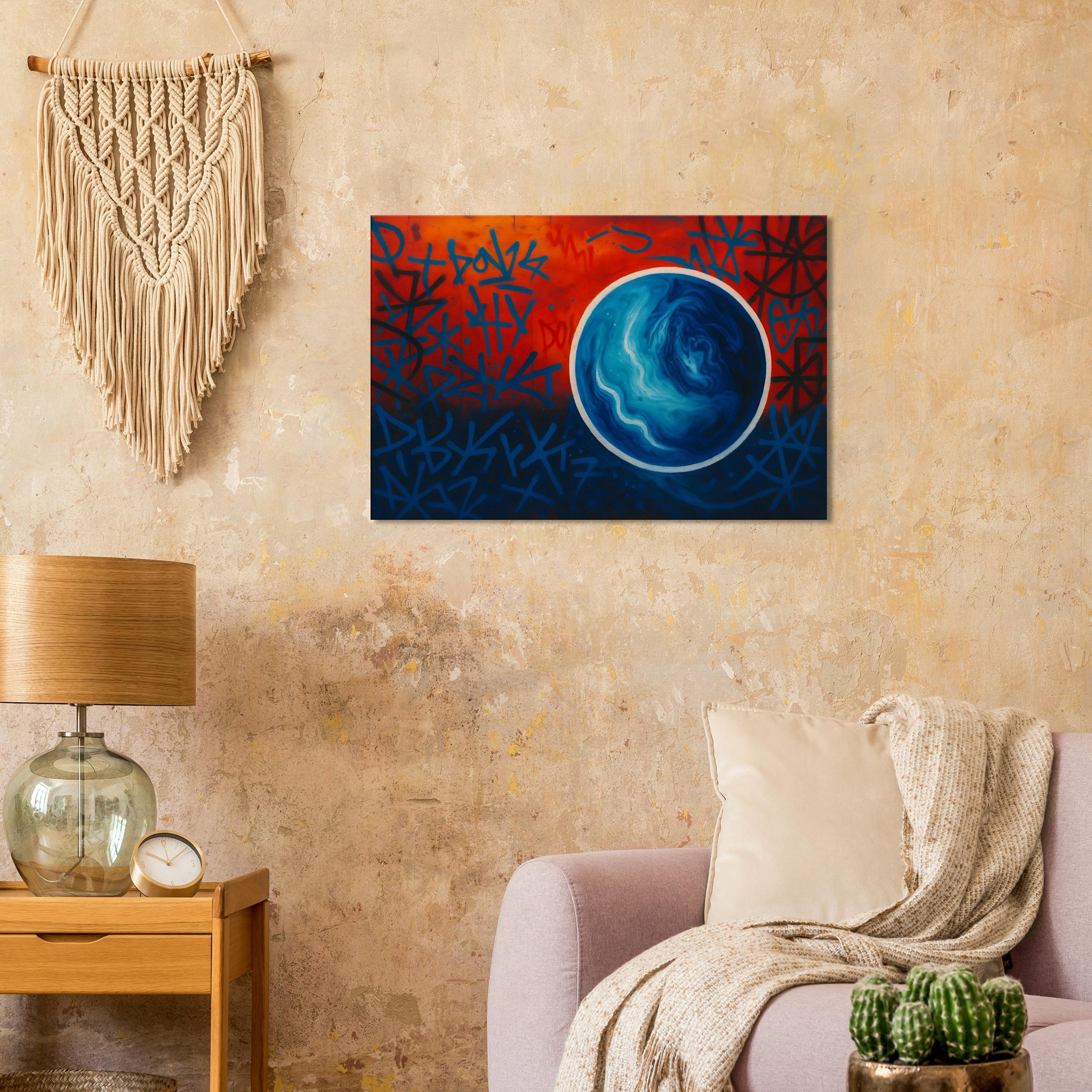 A living room wall with a beige textured wall, a colorful abstract painting with a blue planet, a cream-colored throw blanket on a light pink sofa, a small wooden side table with a glass lamp and a clock, a cactus plant, and a macrame wall hanging.