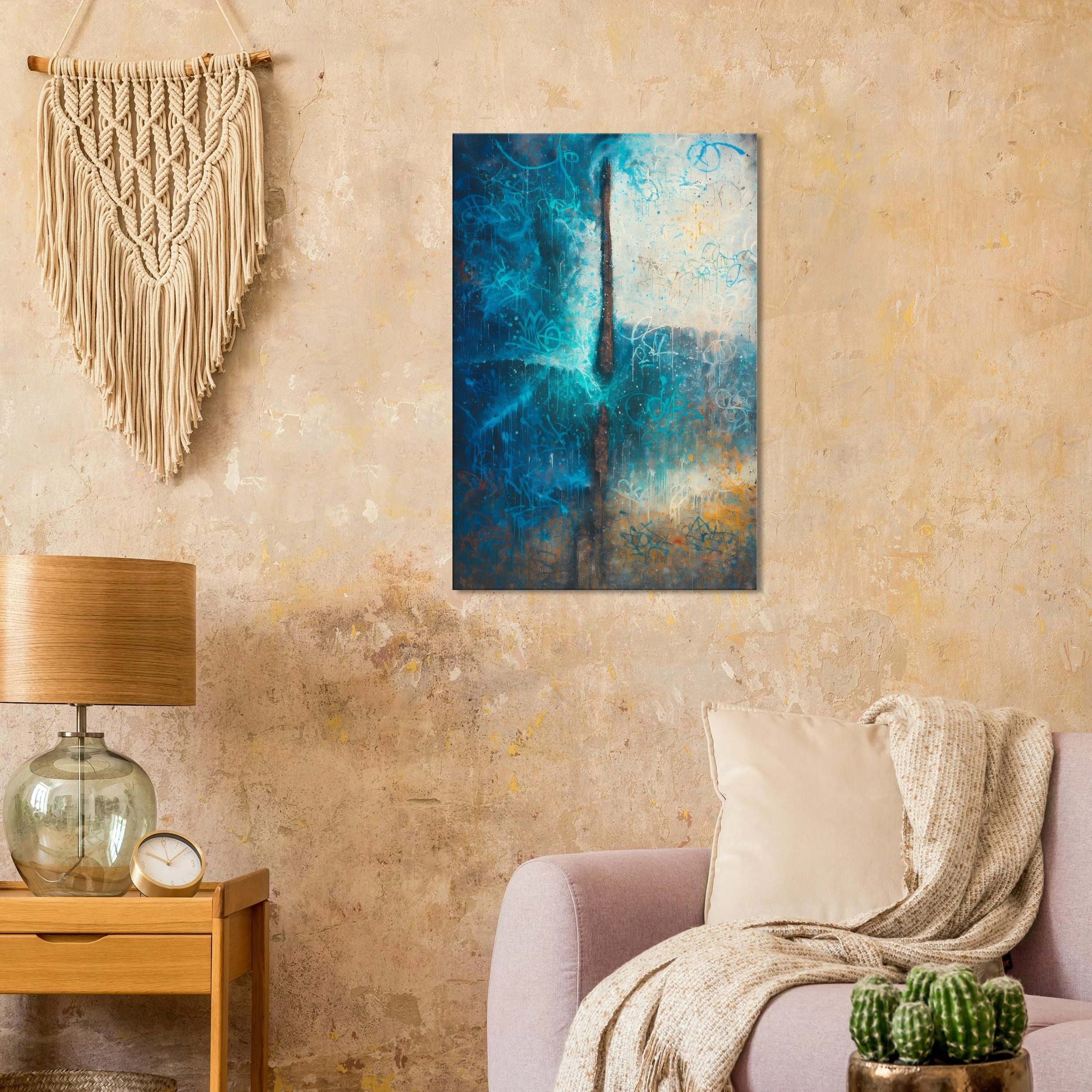 A living room with a textured beige wall, a macramé wall hanging, a modern painting with blue abstract designs, a wooden side table with a glass lamp and a clock, a pale pink couch with a white pillow and beige throw blanket, and a small pot of cacti