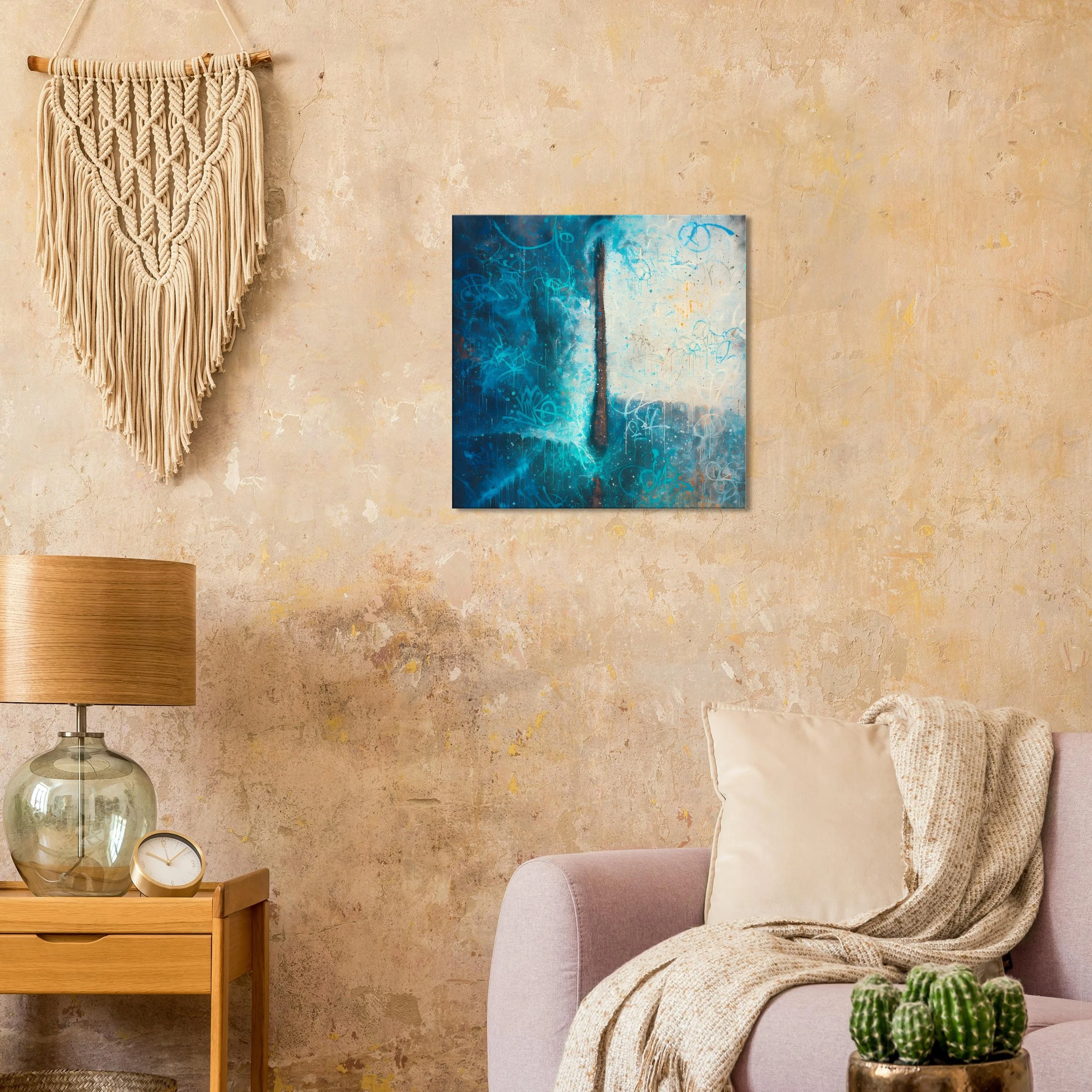 A cozy living room corner with a beige textured wall, a beige armchair with a white pillow and a beige throw blanket, a small wooden side table with a glass lamp and clock, a cactus plant, a wall hanging made of cream-colored macrame, and a blue abstract painting on the wall.