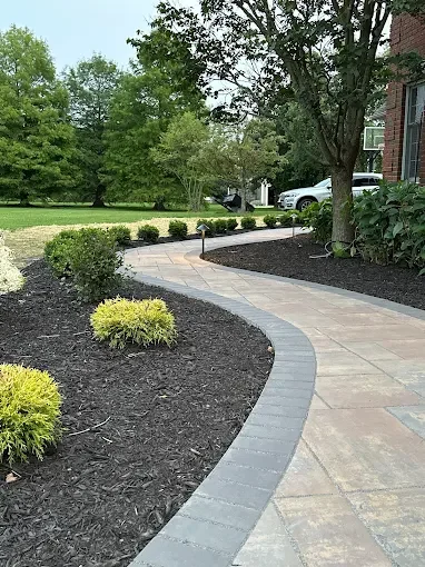 Curved sidewalk with decorative edging, bordered by landscaped garden beds with mulch and small bushes, trees in the background, and part of a residential building on the right.