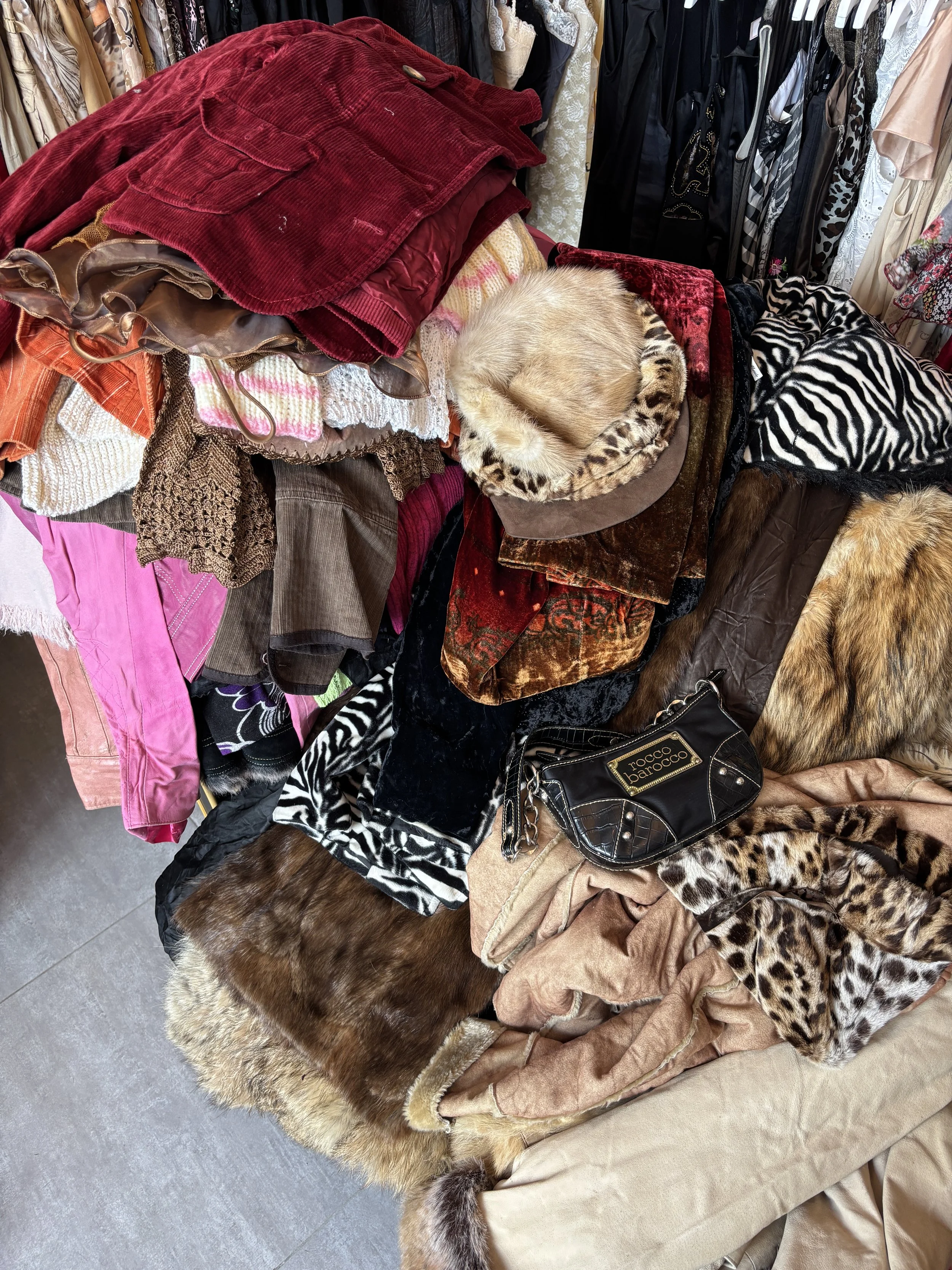 A pile of various clothing items from Dainty Vintage - the vintage store in Malta; including jackets, sweaters, scarves, and fabrics, with a cat curled up sleeping on top of the pile.