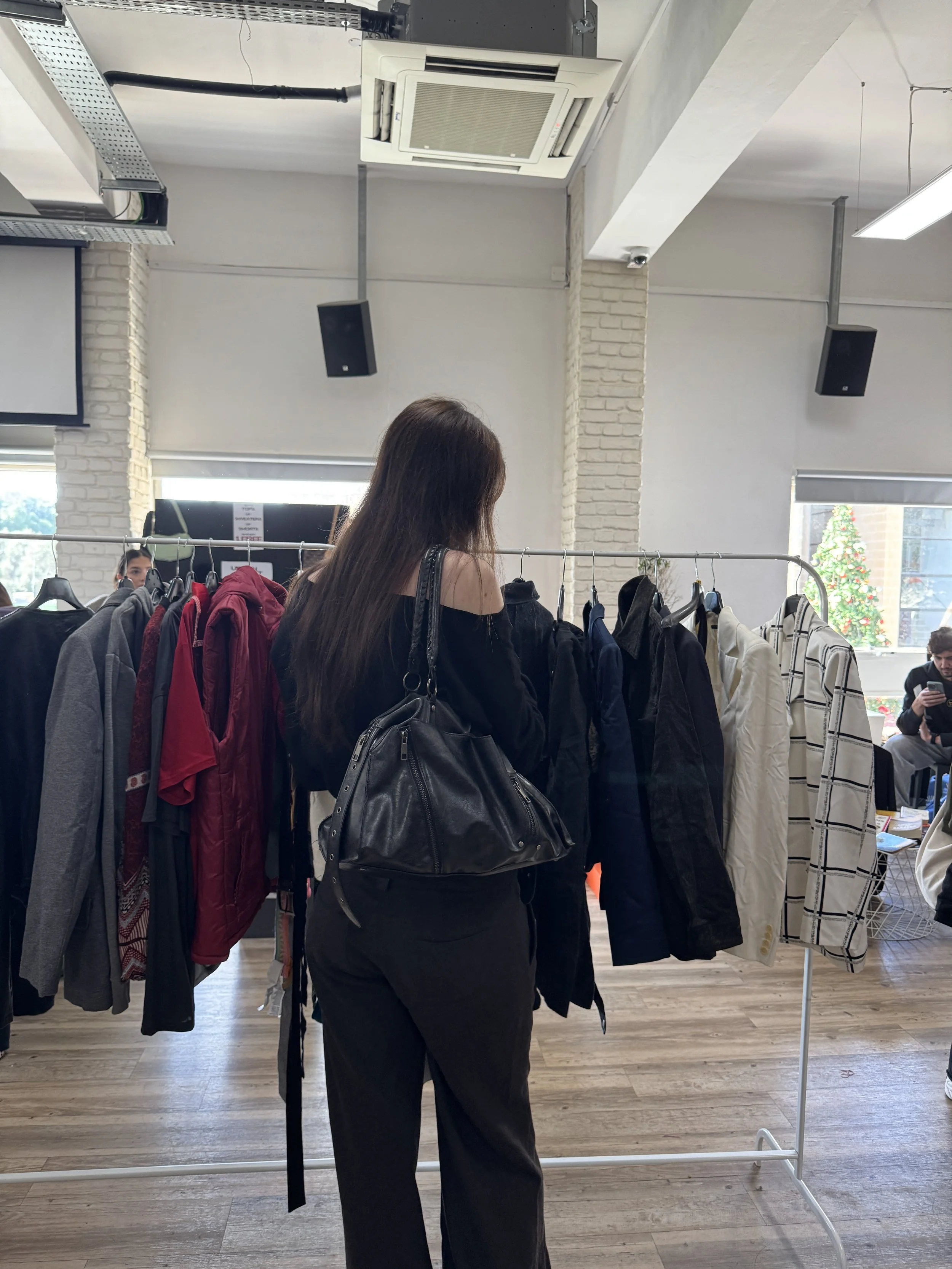 A woman with long hair, dressed in black, shopping at a clothing rack at a Buy the Kilo Event in Malta, with other shoppers visible in the background.