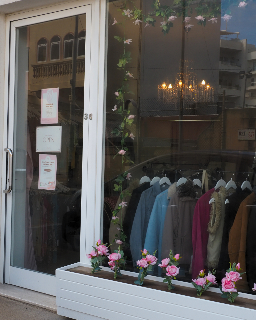 Dainty Vintage's glass window and door displaying clothing inside, with pink flowers in a white planter box outside. Found in Gharghur, Malta.