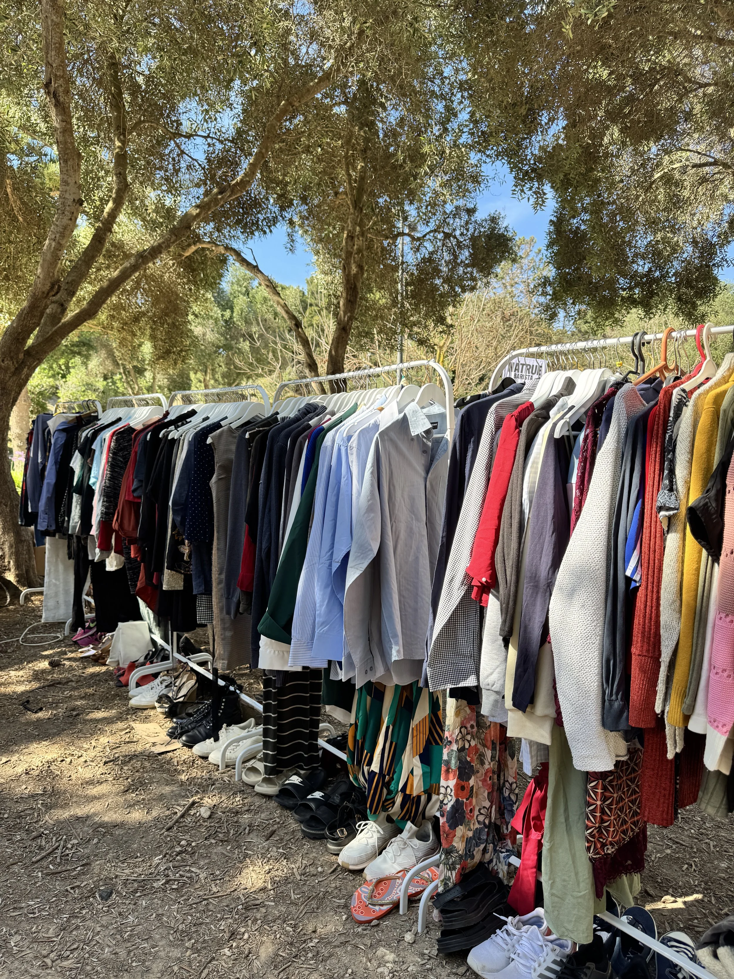 Clothing rack at an outdoor Kilo Sale from Buy the Kilo with various shirts, pants, and shoes under trees on a dirt ground.