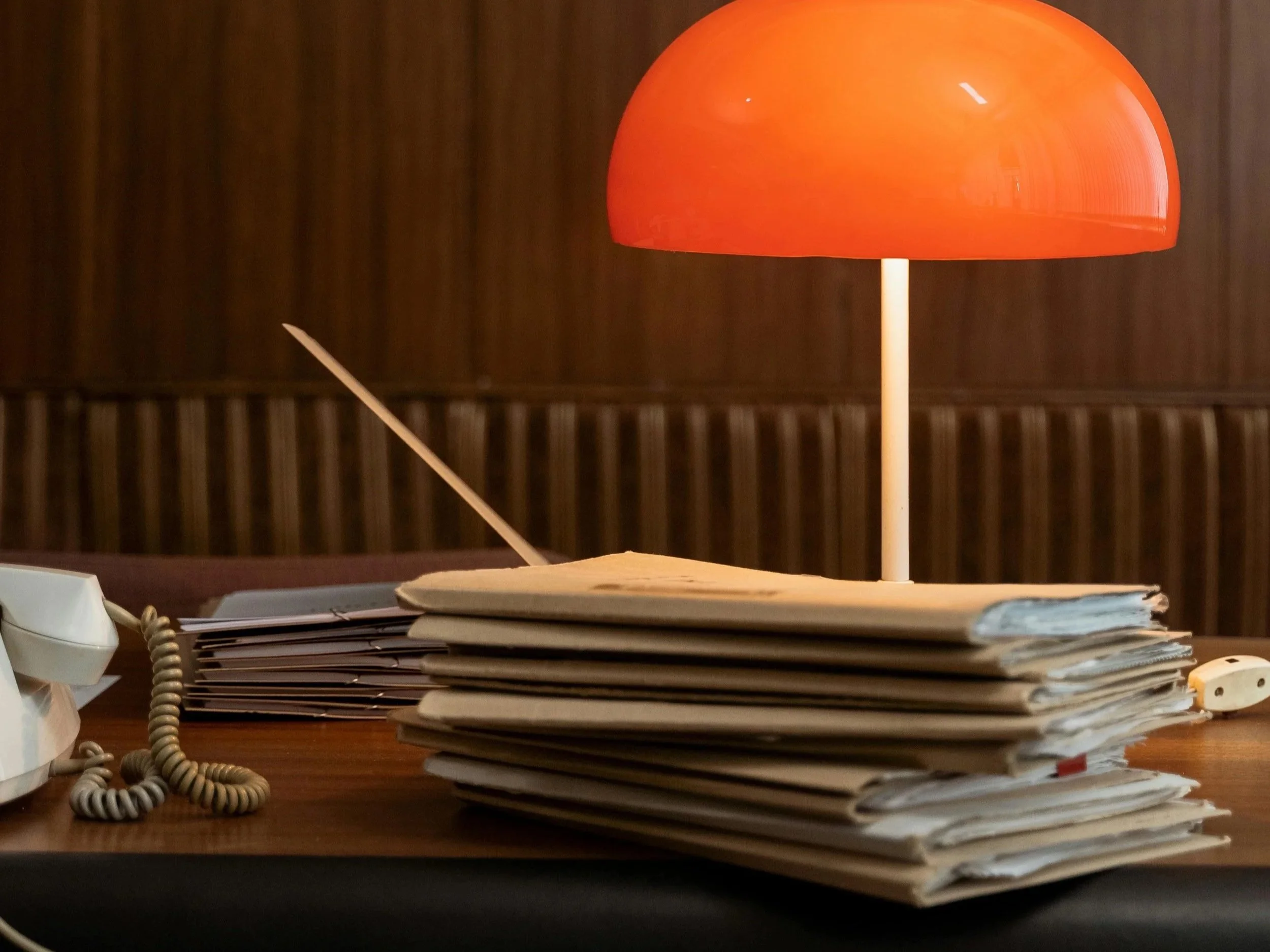 Desk with a large stack of papers, a yellow sticky note on top, an orange table lamp, a vintage phone, and a white power strip.