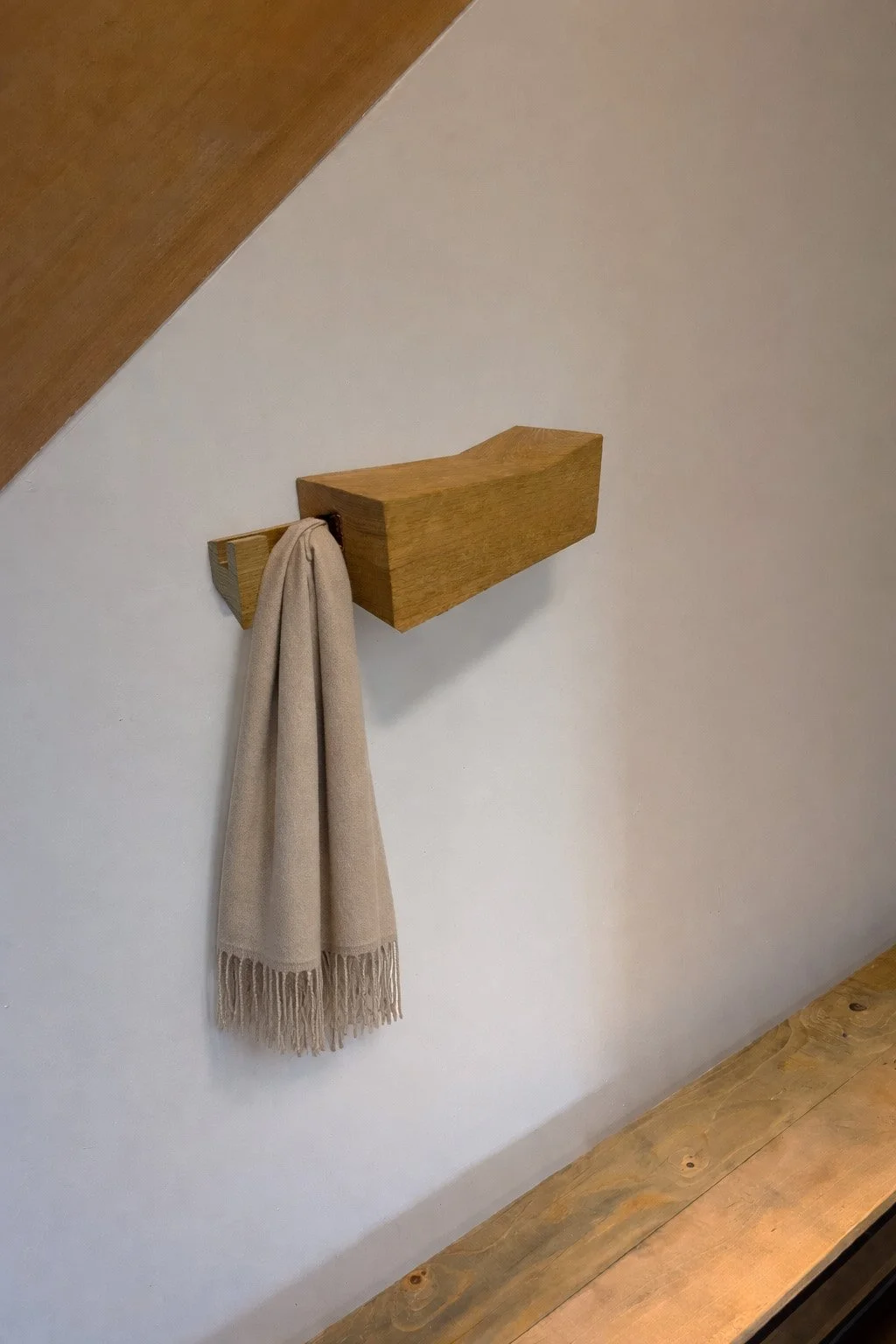 Houten kapstok met een beige sjaal er aan, geplaatst op een witte muur onder een schuine houten plafond.