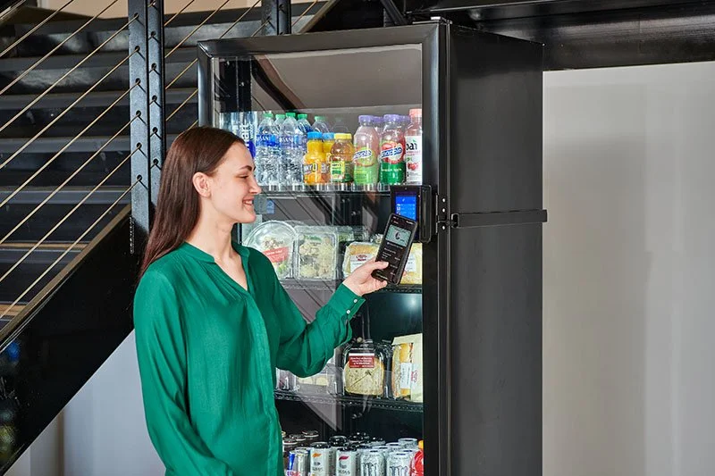 A woman in a green shirt using her phone to interact with a vending machine filled with bottled drinks, packaged snacks, and canned beverages.