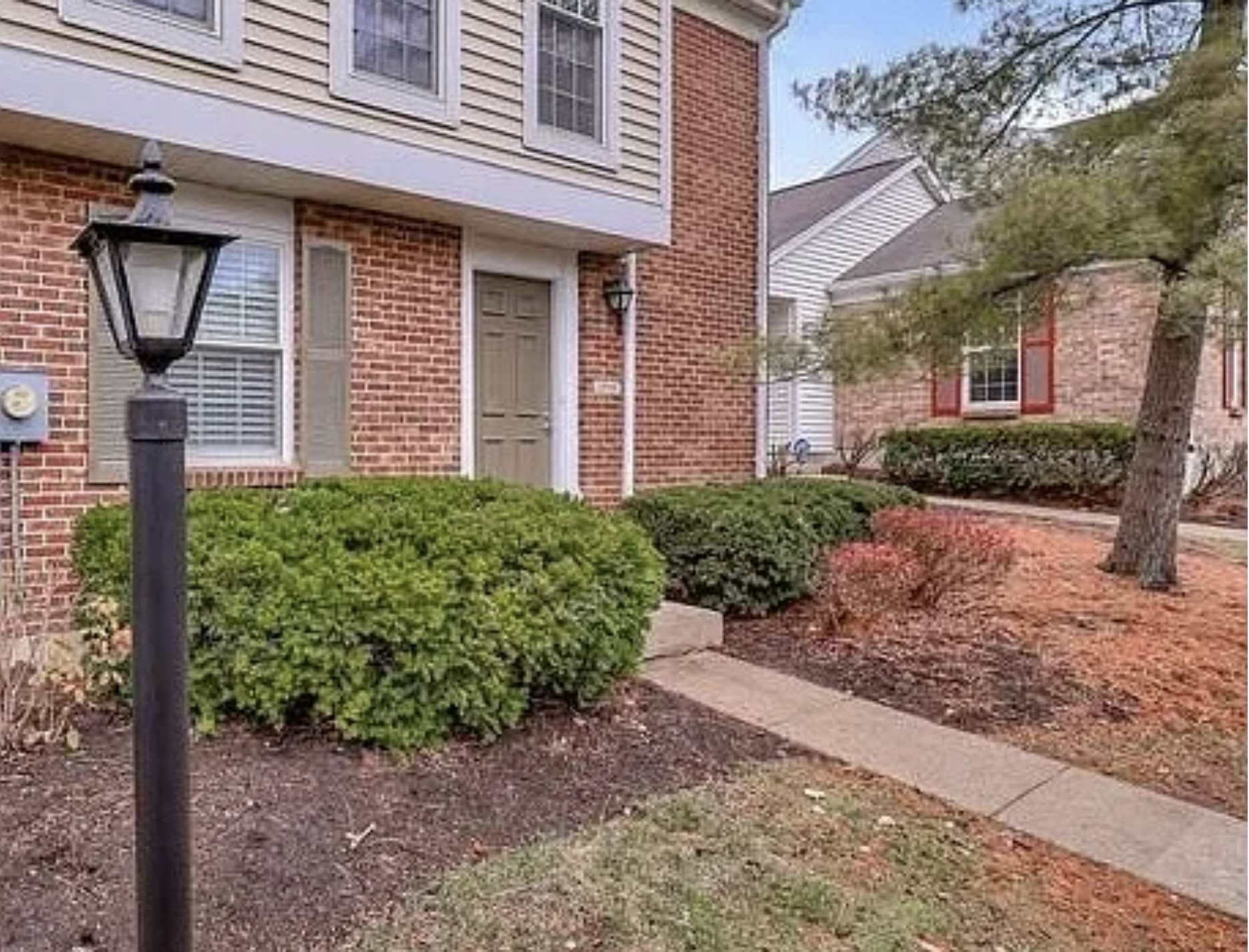 Front yard with bushes, a lamp post, a sidewalk, and a brick house in the background.