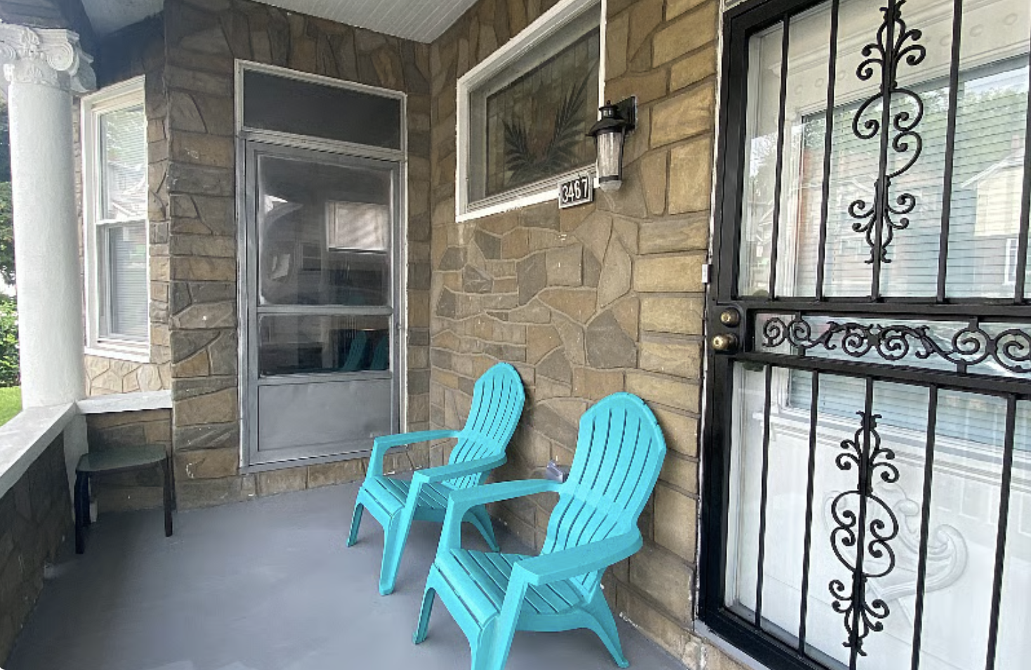 A porch with two blue Adirondack chairs, a small black table, a stone wall, a screen door, a window, and a black iron security door.