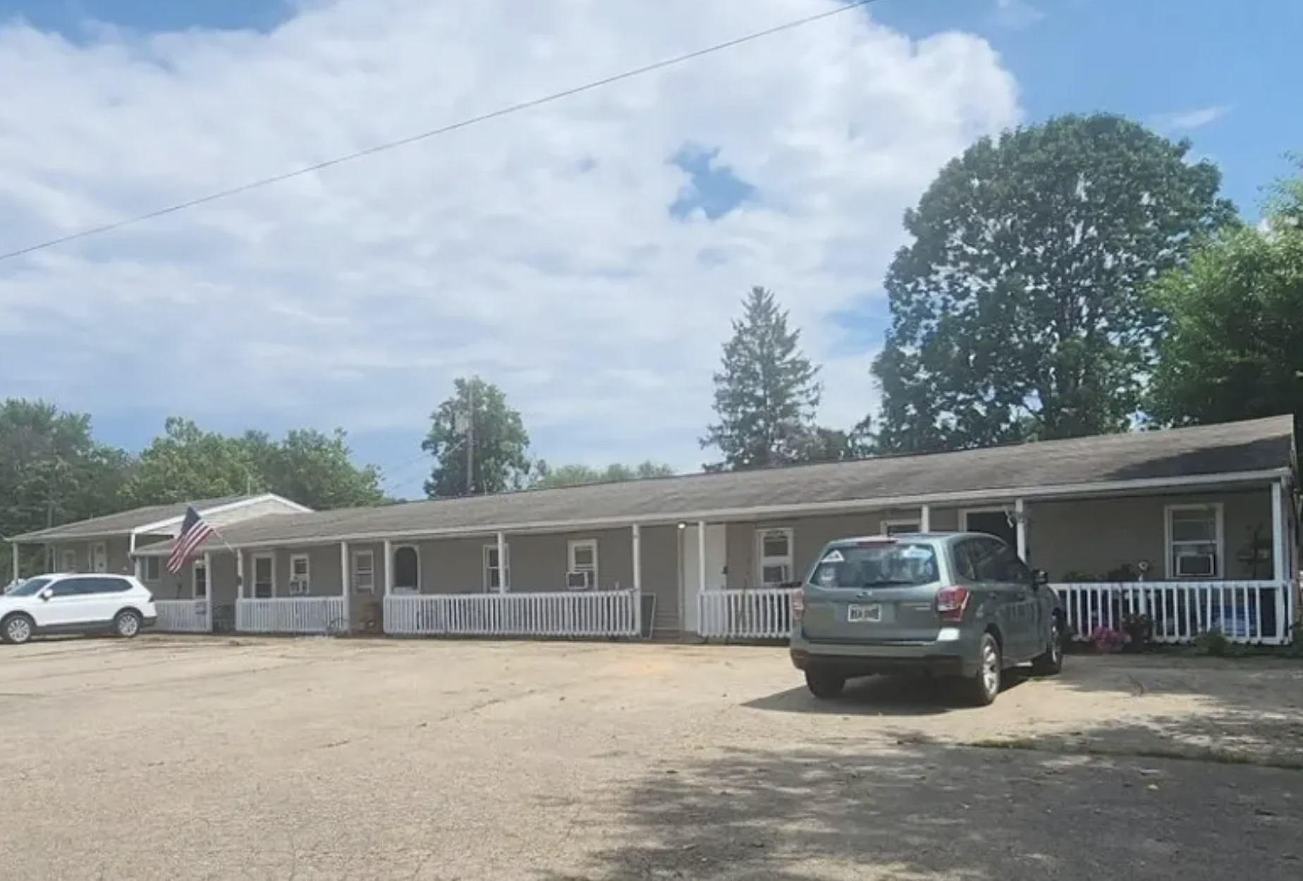 A single-story building with a row of windows and white railings in front. There are two cars parked in the gravel lot, one gray and one white. An American flag is displayed outside the building, and large trees are visible in the background under a partly cloudy sky.