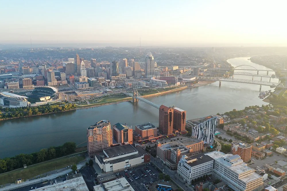 Aerial view of Cincinnati, Ohio, featuring the Ohio River with multiple bridges, downtown skyscrapers, and surrounding cityscape during daylight.