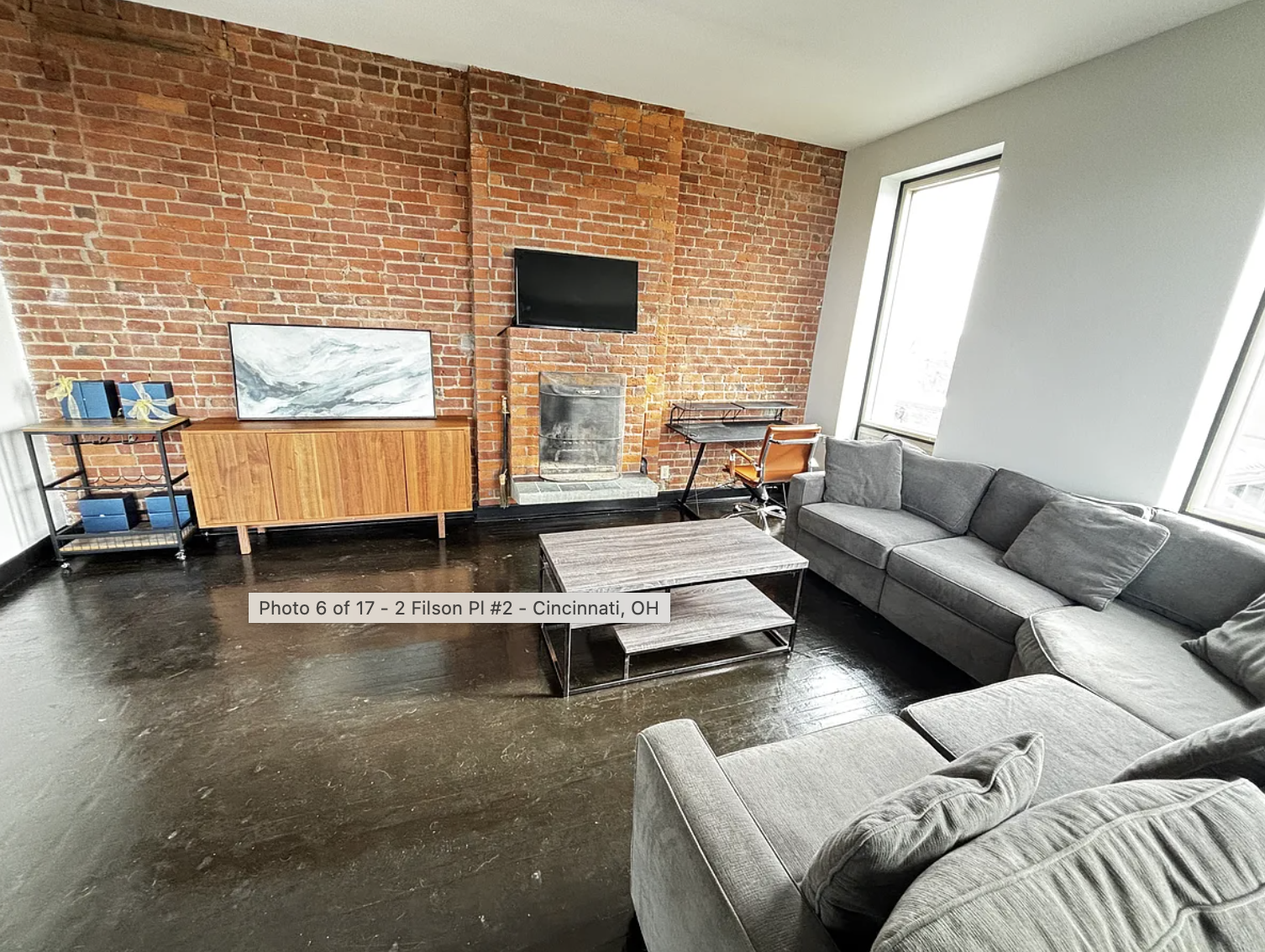 Living room with dark hardwood floors, exposed brick wall, gray sofa, black coffee table, television on brick wall, and large windows.
