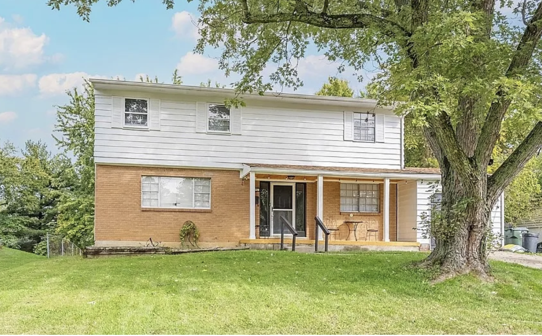 A two-story house with brick and white siding exterior, a front porch with a small table and chairs, a large tree in front, and a well-maintained lawn.