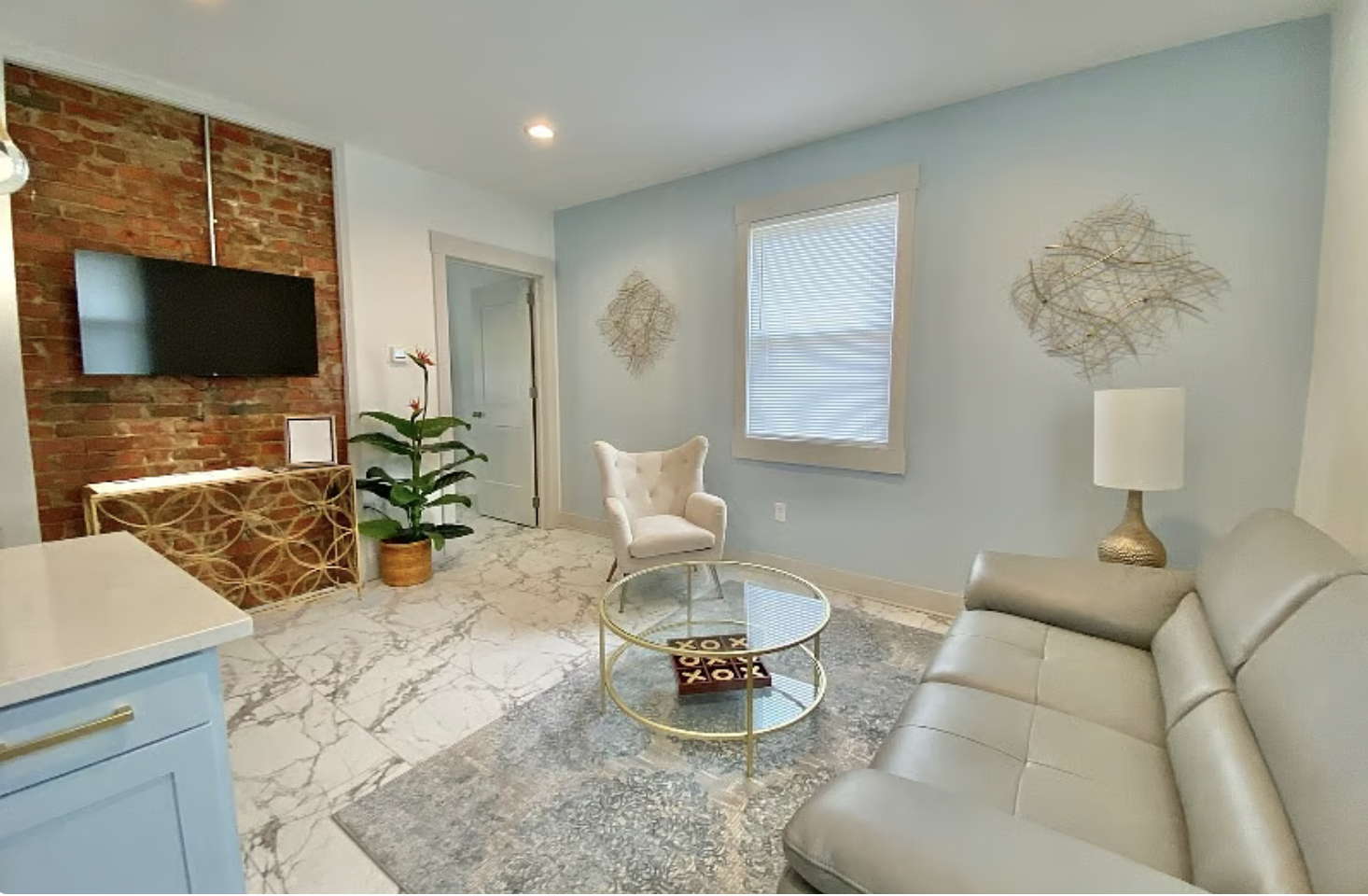 Living room with a light blue wall, a beige leather sofa, a white armchair, a round glass coffee table, a vintage-style wall lamp, a modern lamp on a side table, a black wall-mounted TV on an exposed brick wall, a potted plant, and decorative wall art.