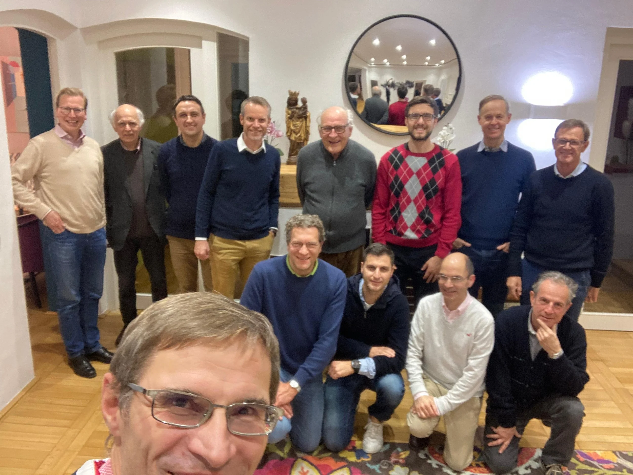 Eine Gruppe von circa 14 Männern, die zusammen in einem Wohnzimmer posieren, einige sitzen, andere stehen, hinter ihnen eine Wand mit Dekorationen, einem runden Spiegel und Kunstgegenständen.