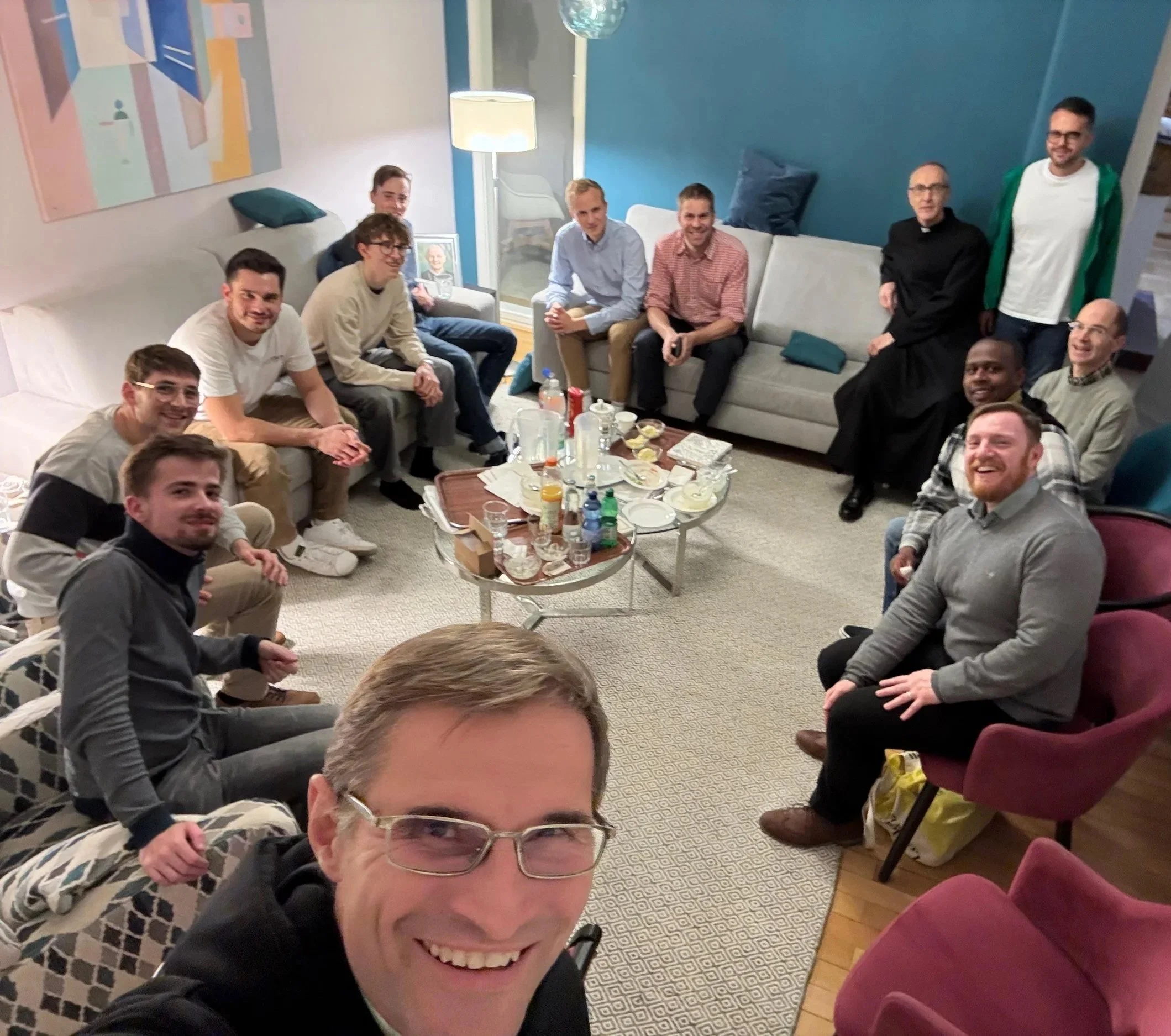 Eine Gruppe von Menschen sitzt in einem Wohnzimmer, einige auf Sofas und Stühlen, während sie für ein Foto lächeln.