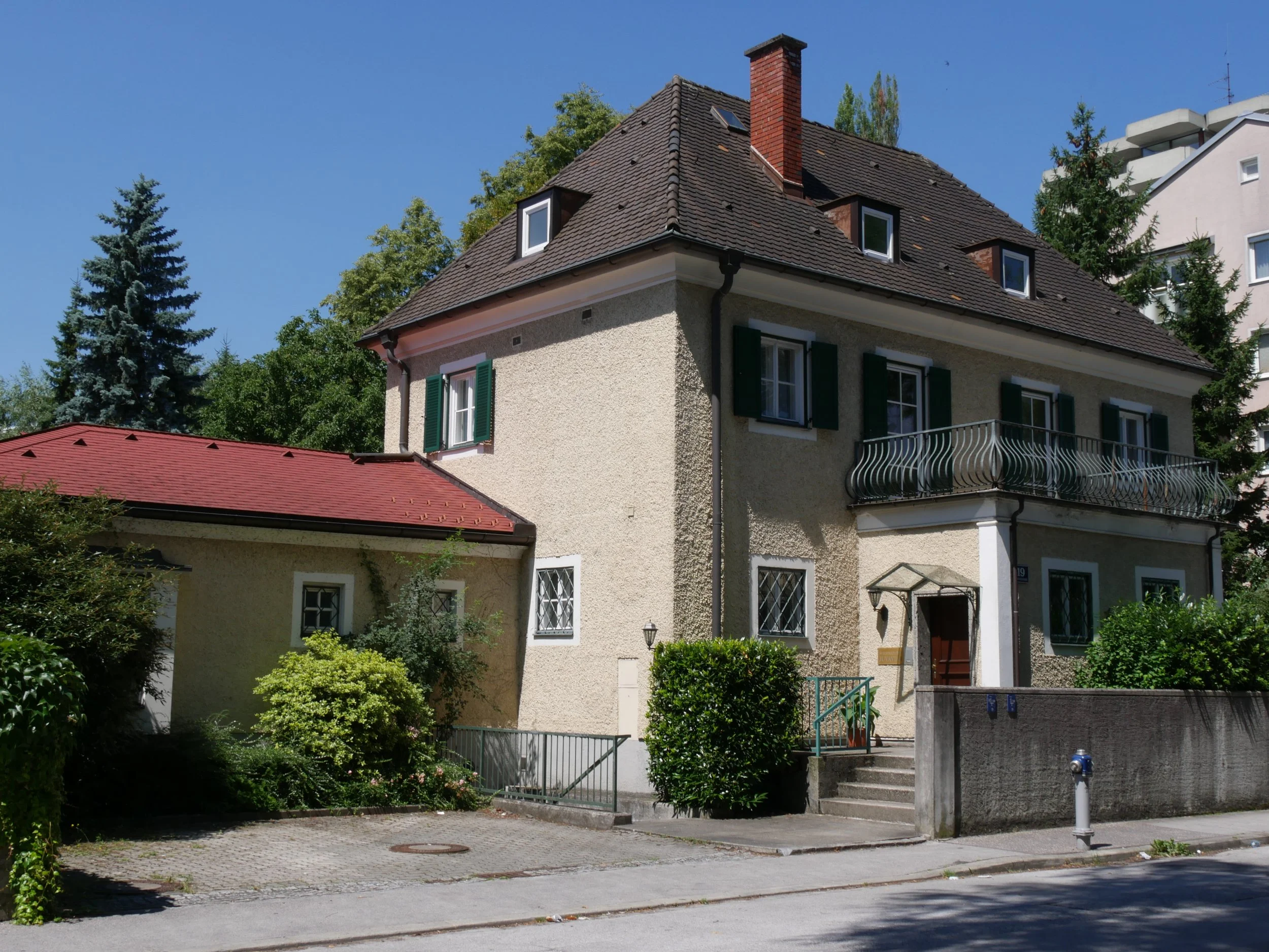 Ein mehrstöckiges Haus mit cremefarbener Fassade, grünen Fensterläden, einer Terrasse mit verzierter Balustrade und einem背eingang, umgeben von Büschen und Bäumen, unter klarem blauen Himmel.