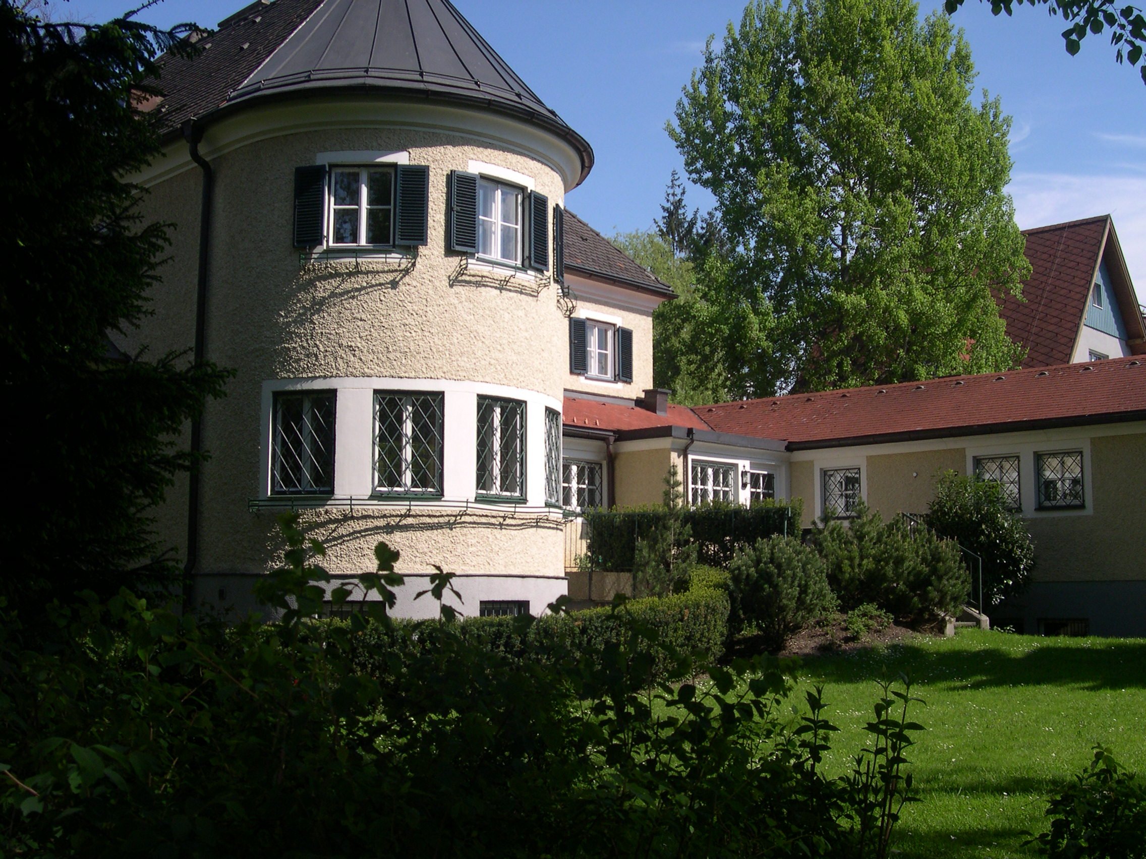 Ein Haus mit runden Ecken und mehreren Fenstern, umgeben von grünen Büschen und Bäumen, bei Sonnenschein.