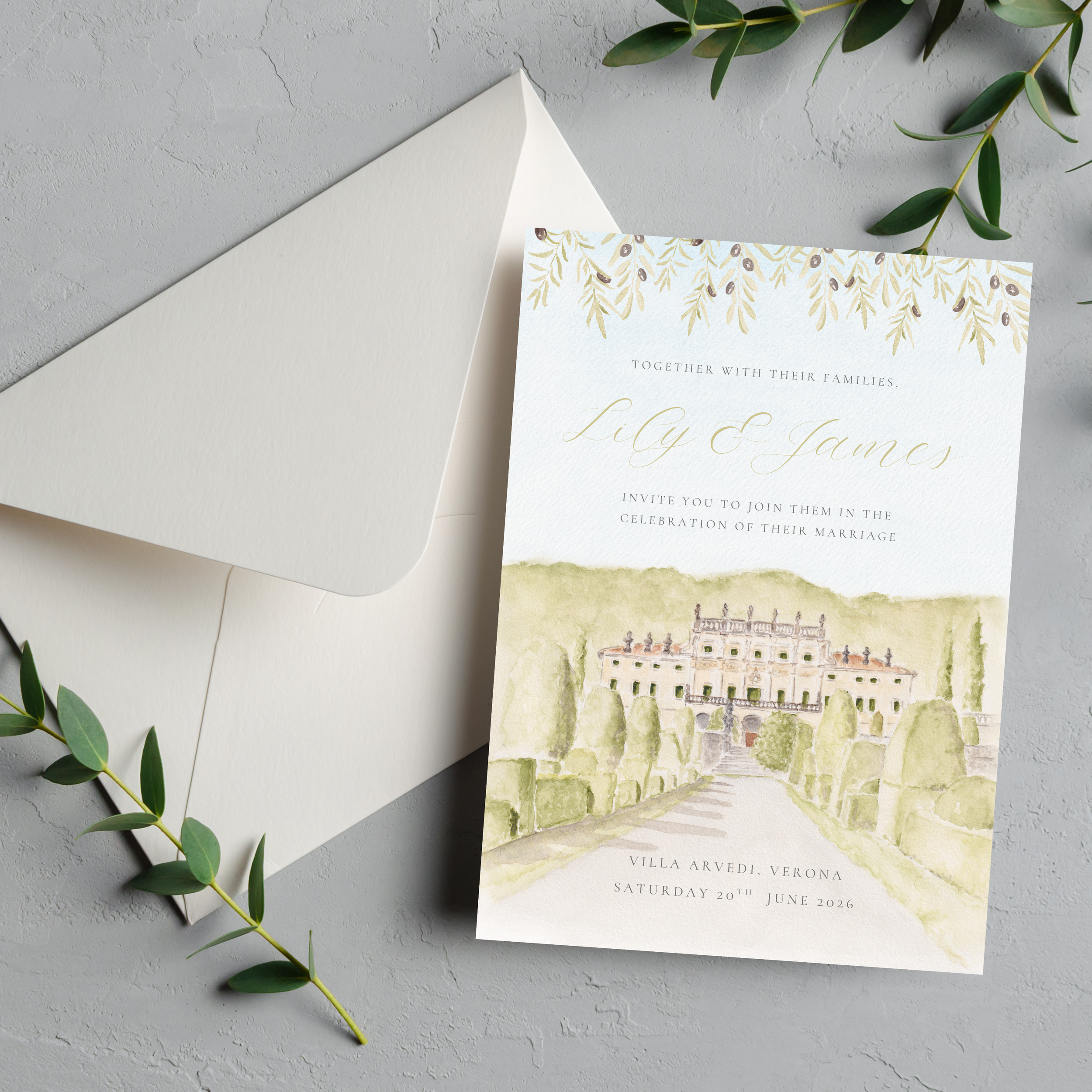 Wedding invitation card featuring a watercolor illustration of a villa with greenery, placed on a light grey surface with an envelope and green leaves beside it.