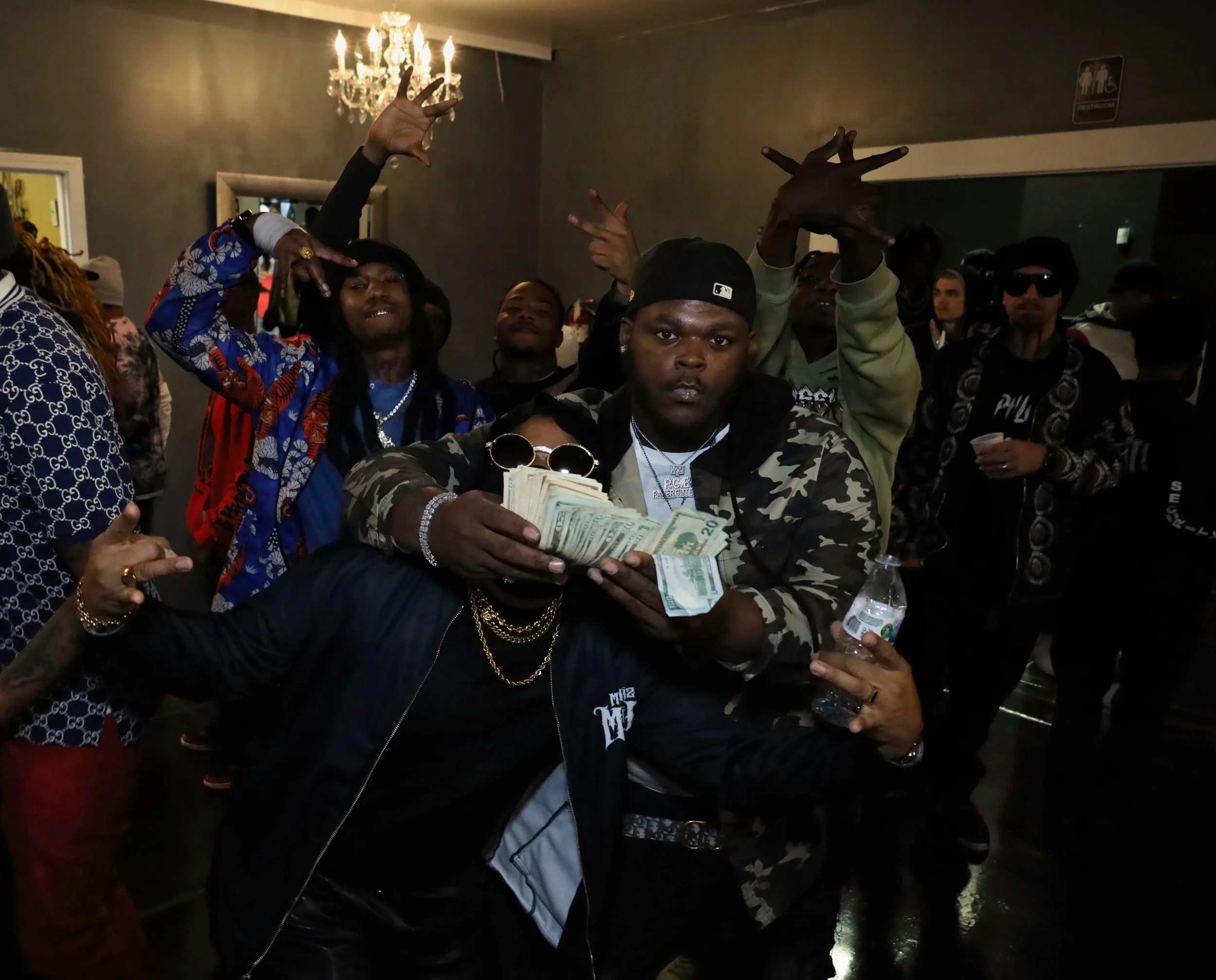A group of young men and women gathered at a party or club, with one person in the front holding a stack of cash and others making hand gestures. The setting appears to be indoors with a chandelier overhead.
