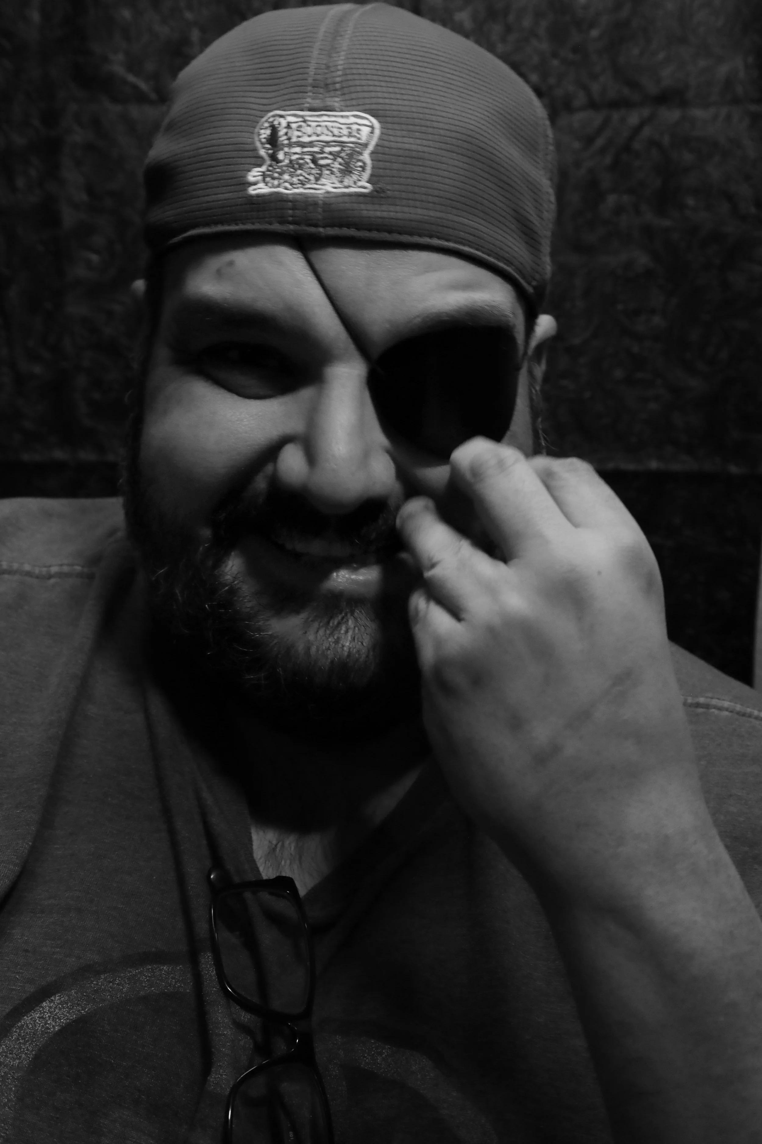 A man with a beard wearing a cap and glasses hanging from his shirt, holding an eye patch over one eye, smiling at the camera, in a black and white photo.
